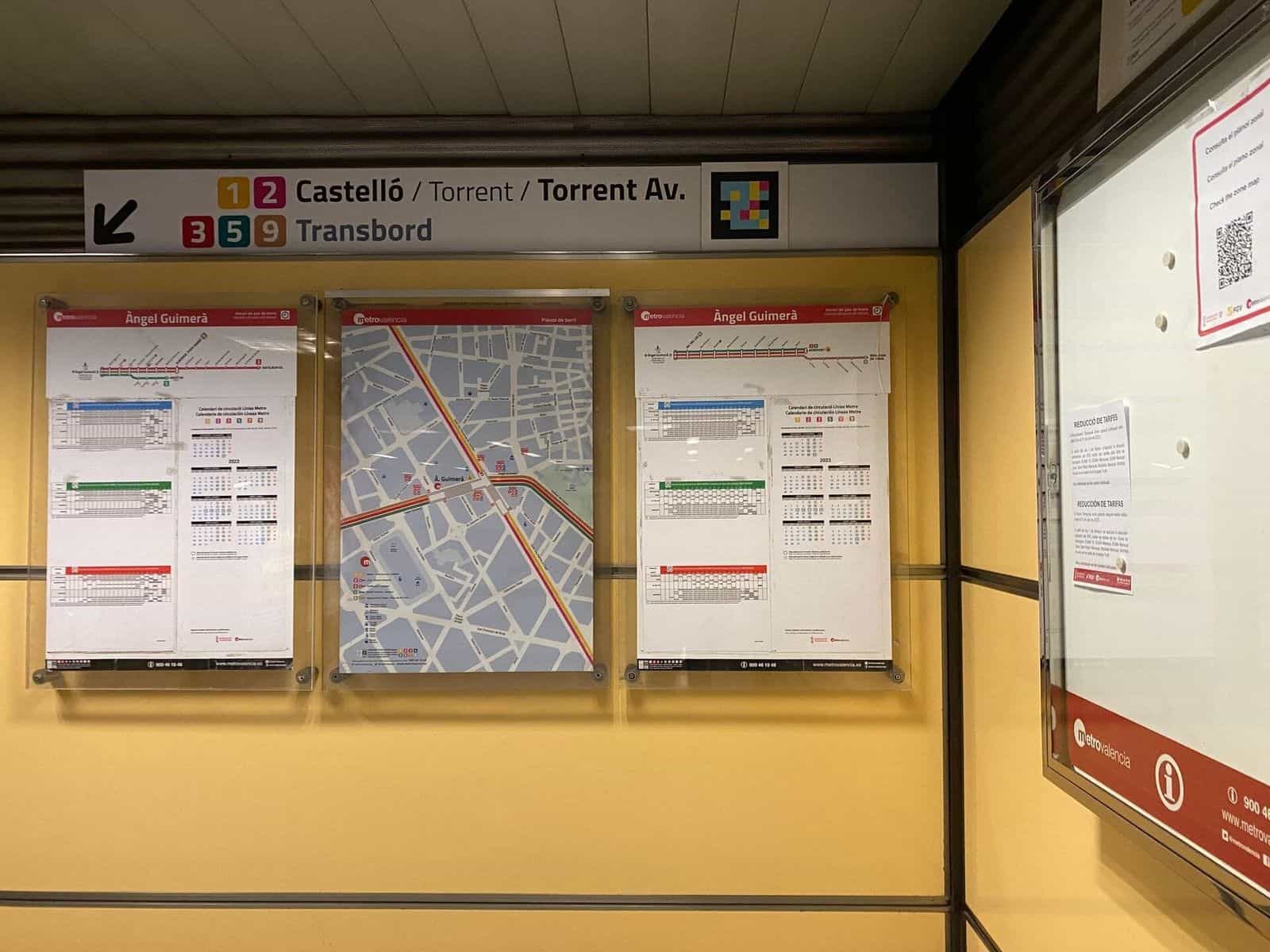 Pannello direzionale di cambio del mezzanino con l’indicazione «1 2 Castelló / Torrent / Torrent Av.» e sotto «3 5 9 Transbord»; accanto al cartello, un codice NaviLens quadrato a colori e, sulla parete inferiore, le piante di zona e il calendario di circolazione di Àngel Guimerà