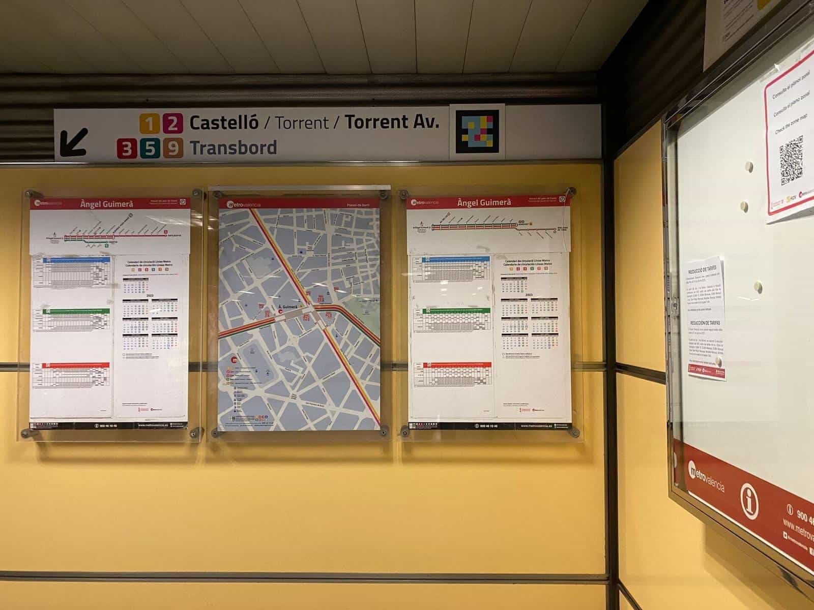 Mezzanine interchange sign reading '1 2 Castelló / Torrent / Torrent Av.' and below '3 5 9 Transbord'; next to the sign, a colourful square NaviLens code and, on the wall below, the zonal maps and circulation calendar of Àngel Guimerà.