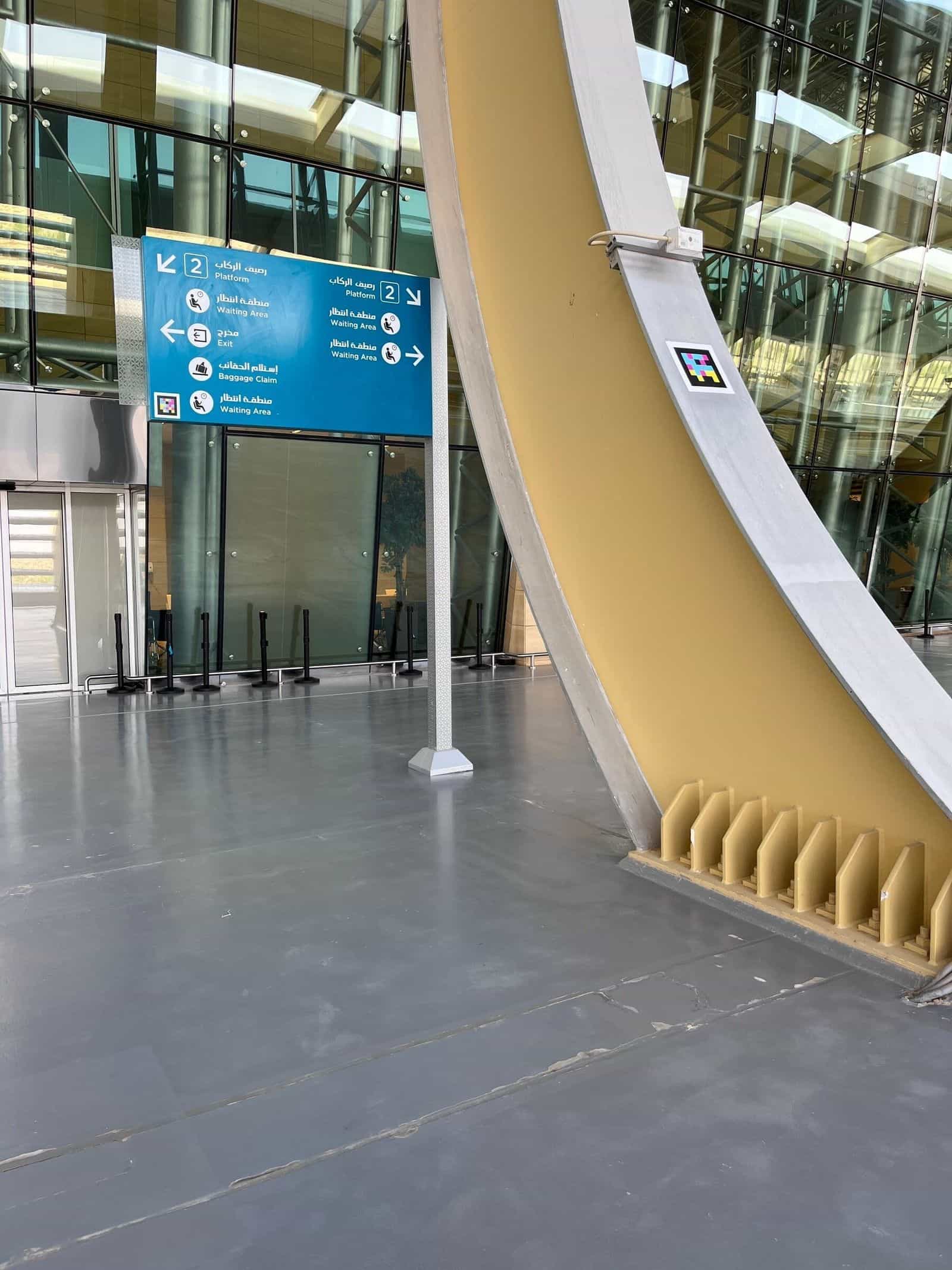 「رصيف الركاب / Platform 2」「Waiting Area」「Exit」「Baggage Claim」への矢印が示された青いSAR方向案内板。横の黄色いアーチにはNaviLensコード