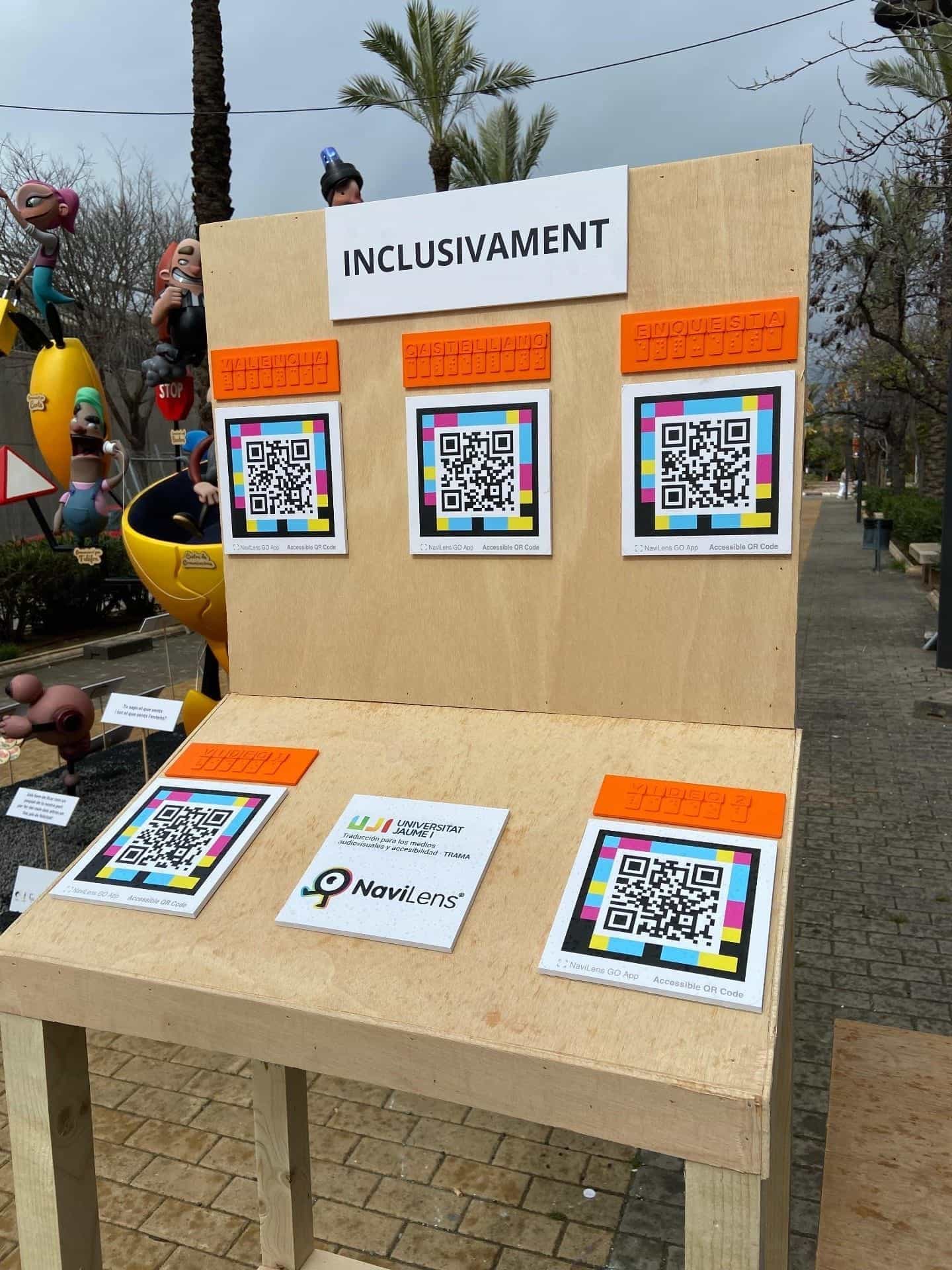 Dettaglio del pannello «INCLUSIVAMENT»: tre codici NaviLens quadrati a colori di grande formato sotto etichette arancioni in braille con le diciture «VALÈNCIA», «CASTELLANO» ed «ENQUESTA»; alla base altri due codici con le diciture «VÍDEO 1» e «VÍDEO 2» e, al centro, la targa con i logo di UJI · TRAMA e NaviLens