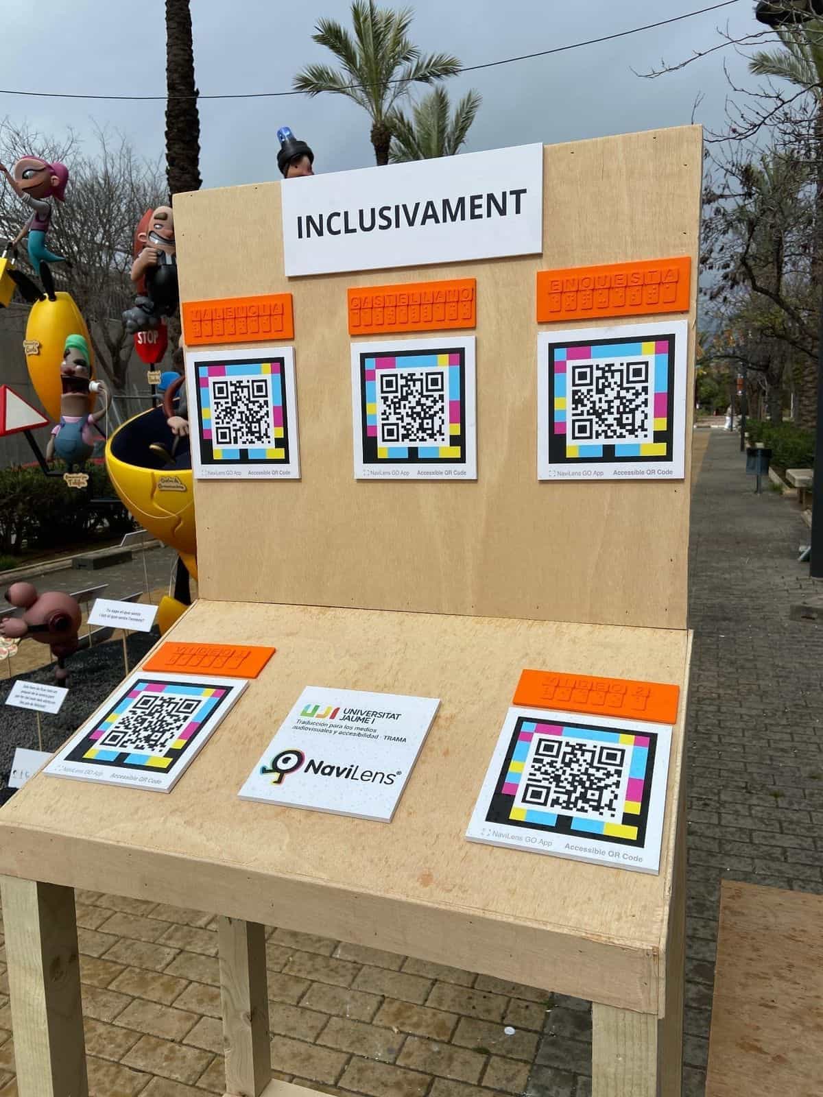 Detalle del panel ‘INCLUSIVAMENT’: tres códigos NaviLens cuadrados a colores de gran formato bajo etiquetas naranjas en braille con los rótulos «VALÈNCIA», «CASTELLANO» y «ENQUESTA»; en la base, otros dos códigos rotulados «VÍDEO 1» y «VÍDEO 2», y al centro la placa con los logos de UJI · TRAMA y NaviLens