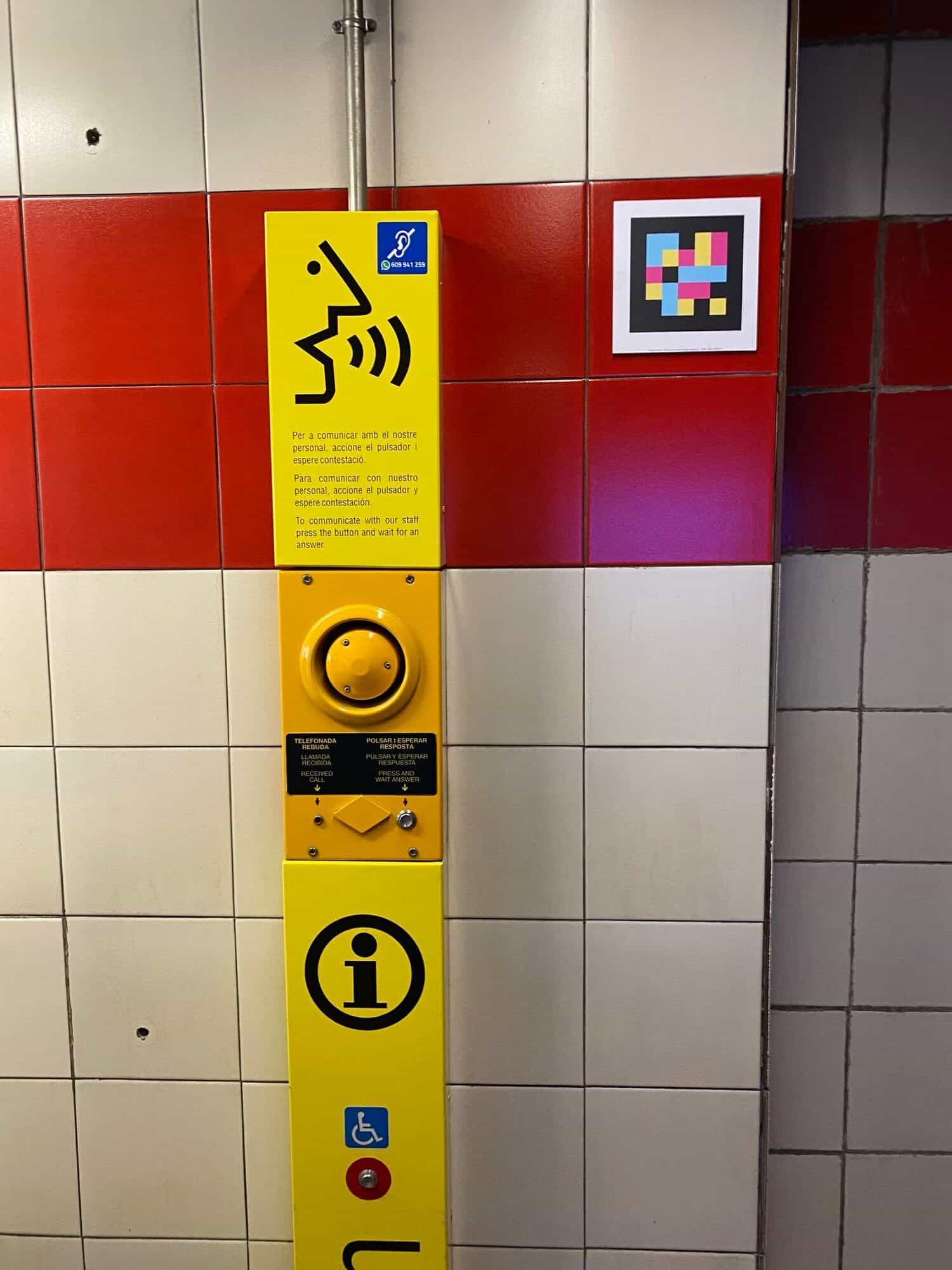 Pannello interfono giallo sulla parete dell’atrio con altoparlante circolare, pulsante a forma di diamante e testi trilingui in valenciano, spagnolo e inglese: «POLSAR I ESPERAR RESPOSTA / PULSAR Y ESPERAR / PRESS AND WAIT ANSWER», accanto a una parete rivestita di piastrelle rosso intenso, viola e bianche