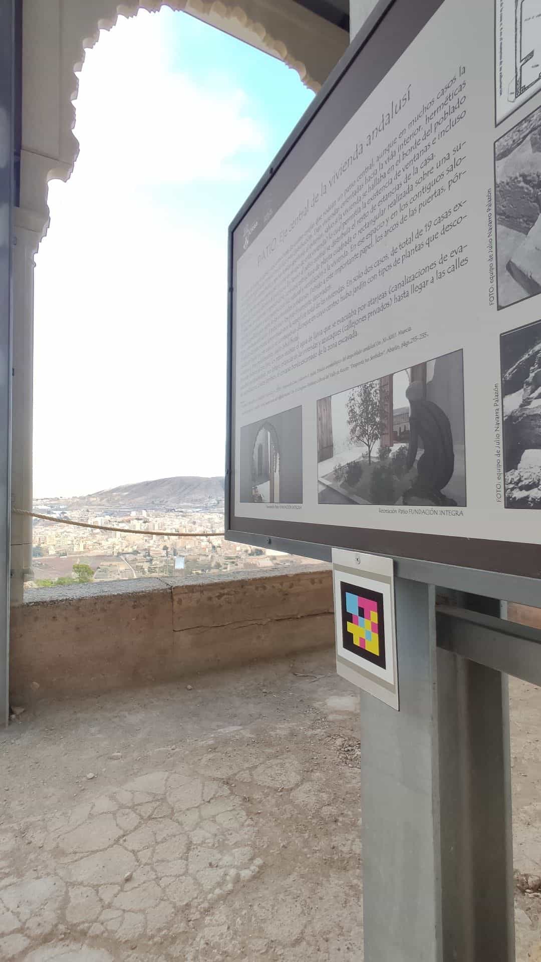 Panel «Patio · eje central de la vivienda andalusí» de la Medina Siyâsa junto a un arco lobulado restaurado, con código NaviLens en el poste