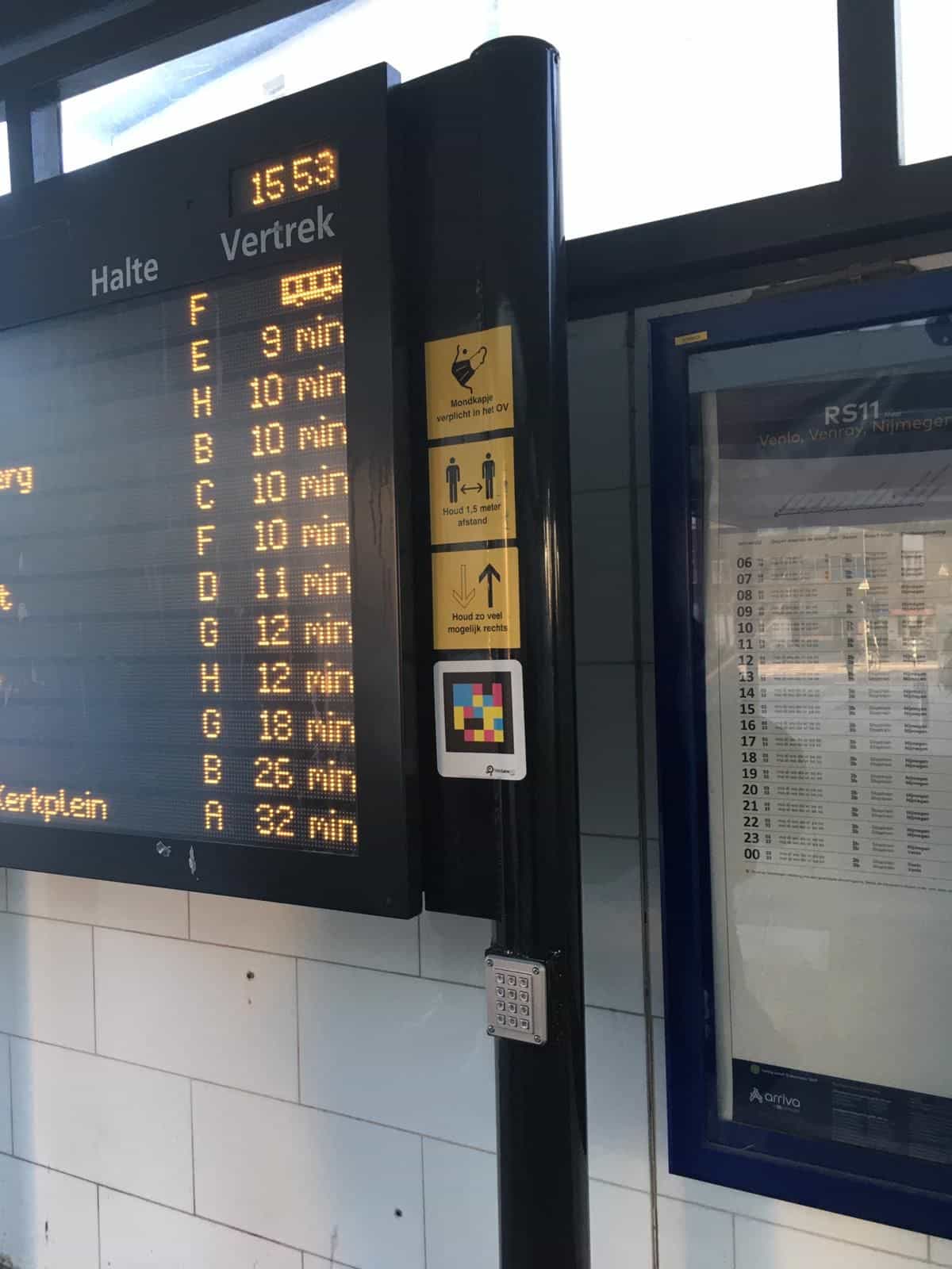 Panel de salidas Halte/Vertrek del busstation Roermond junto a un código NaviLens y al horario impreso de la línea RS11 de Arriva hacia Venlo, Venray y Nijmegen