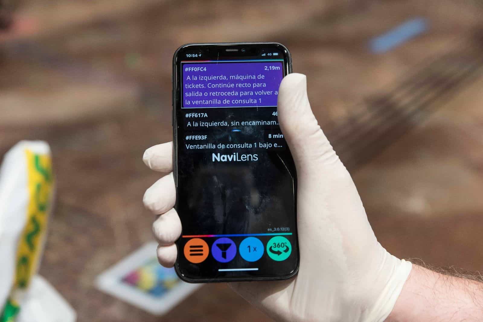 Dettaglio dello schermo di un iPhone con l’app NaviLens che rileva più codici nella hall del Comune di Saragozza: il codice #FF0FC4 a 2,19 m annuncia «A sinistra, biglietteria automatica. Prosegua dritto per l’uscita o torni indietro per rientrare allo sportello informazioni 1», con altri codici a 46 cm e a 8 minuti di distanza