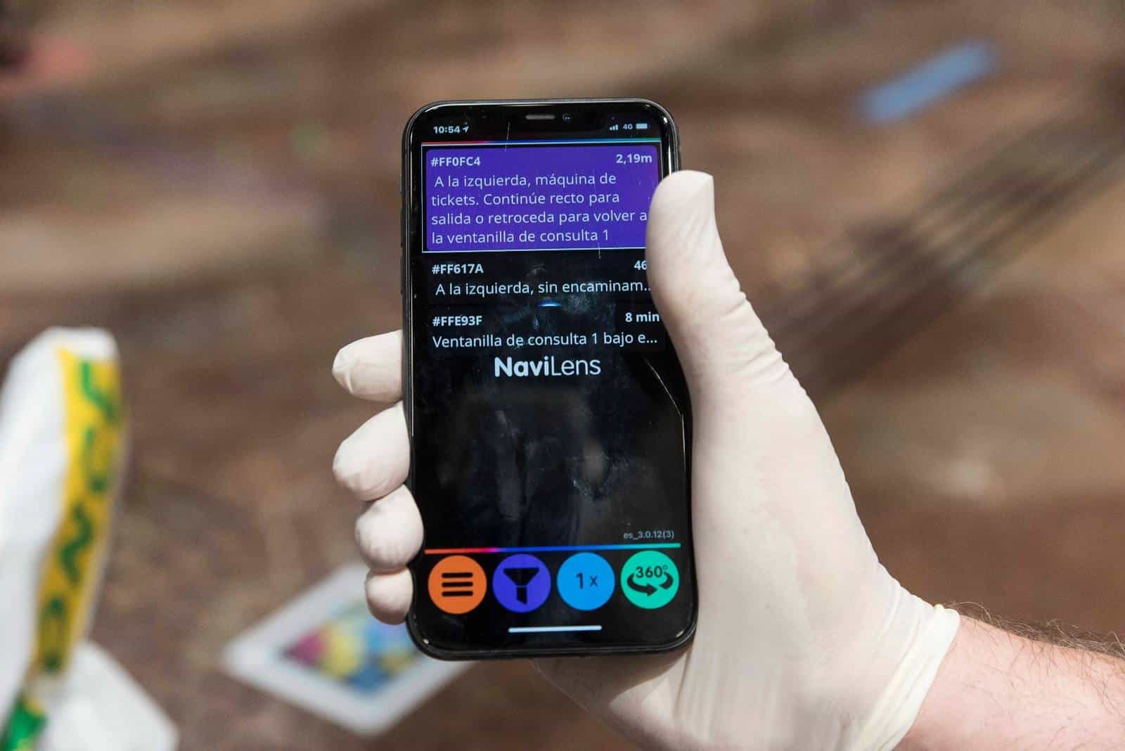 Close-up of an iPhone screen with the NaviLens app detecting several codes in the foyer of Zaragoza City Hall: code #FF0FC4, 2.19 m away, announces «A la izquierda, máquina de tickets. Continúe recto para salida o retroceda para volver a la ventanilla de consulta 1», with other codes 46 cm and 8 min away