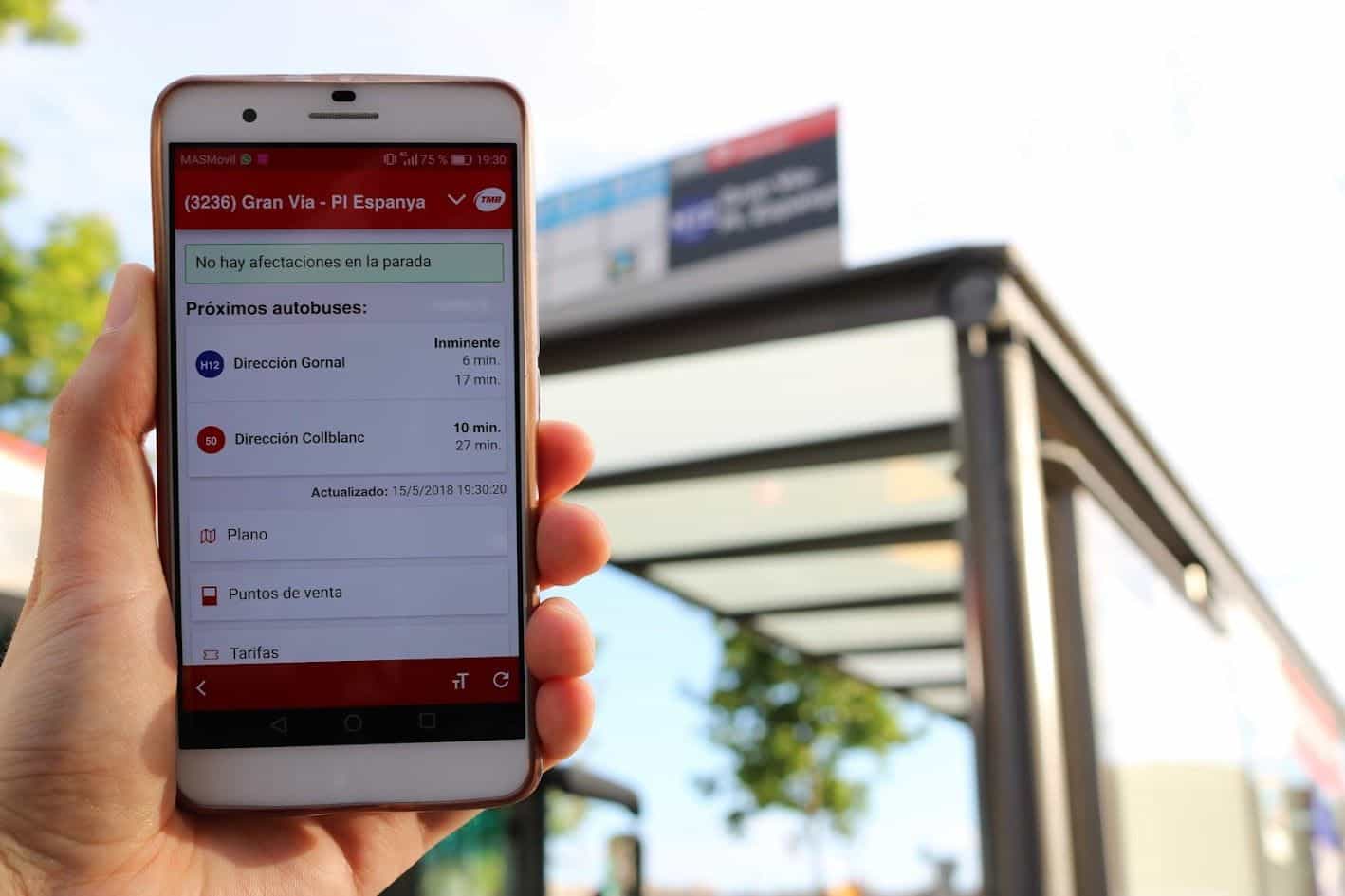 Mano che regge un telefono con l’app NaviLens GO alla fermata (3236) Gran Via — Pl Espanya, con i prossimi autobus H12 e 50, davanti a una pensilina con pubblicità.