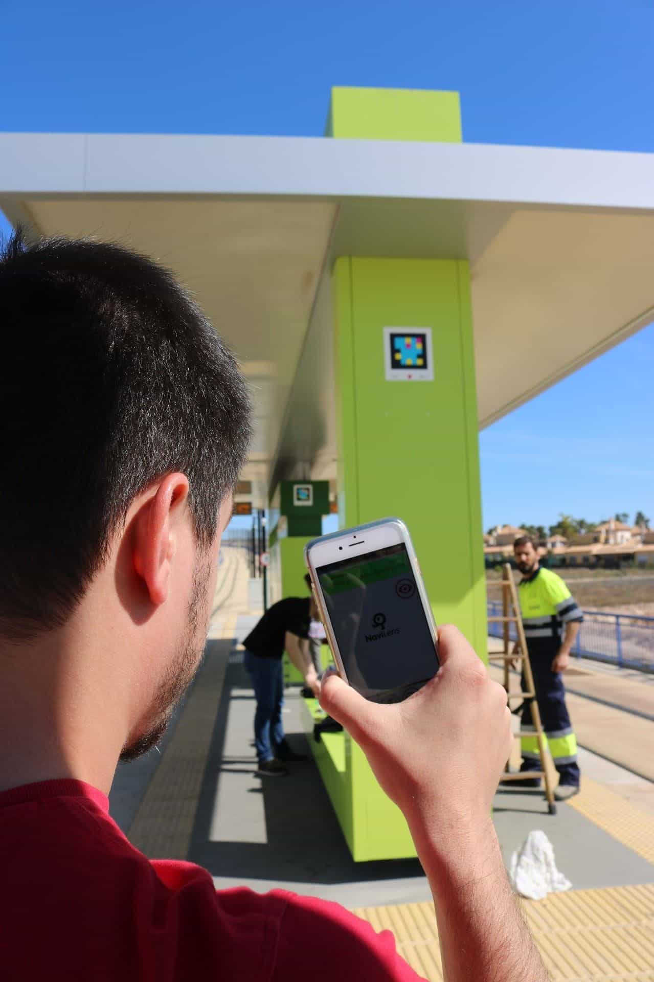 Persona che inquadra un codice NaviLens collocato in cima a una pensilina del Tranvía de Murcia, con lo schermo LED dei prossimi arrivi in fondo