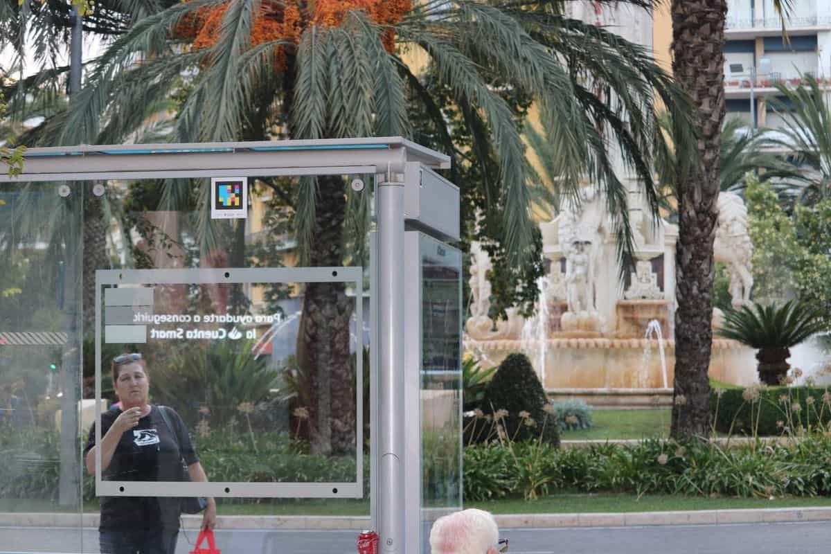 Bus shelter at Plaza de los Luceros in Alicante with a NaviLens code on the canopy and the monumental fountain in the background