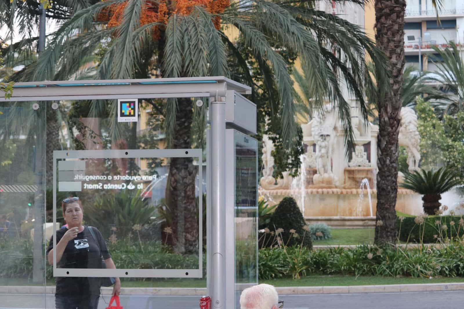 Bus shelter at Plaza de los Luceros in Alicante with a NaviLens code on the canopy and the monumental fountain in the background