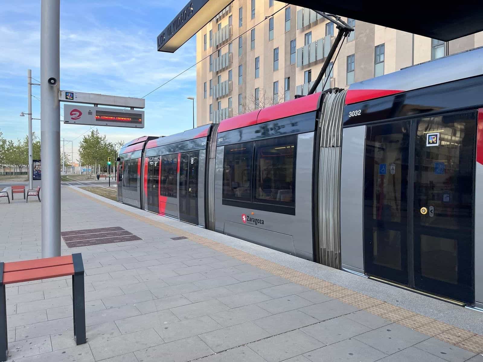 Fermata «Los Olvidados» del tram di Saragozza al tramonto: tram grigio con fascia rossa di Avanza Zaragoza, numero 3032/3033/3034, fermo in banchina, pensilina con l’insegna «Los Olvidados» e codice NaviLens applicato in cima alla palina della fermata e un altro su una delle porte del tram