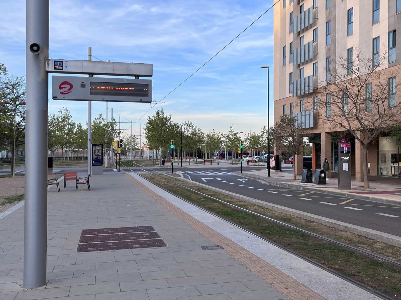 Fermata del tram di Saragozza con pannello led dei tempi di attesa di Avanza con il logo rosso e un codice NaviLens applicato sopra il pannello; sullo sfondo, semafori verdi, binari del tram e un totem metallico del bus dall’altro lato del viale