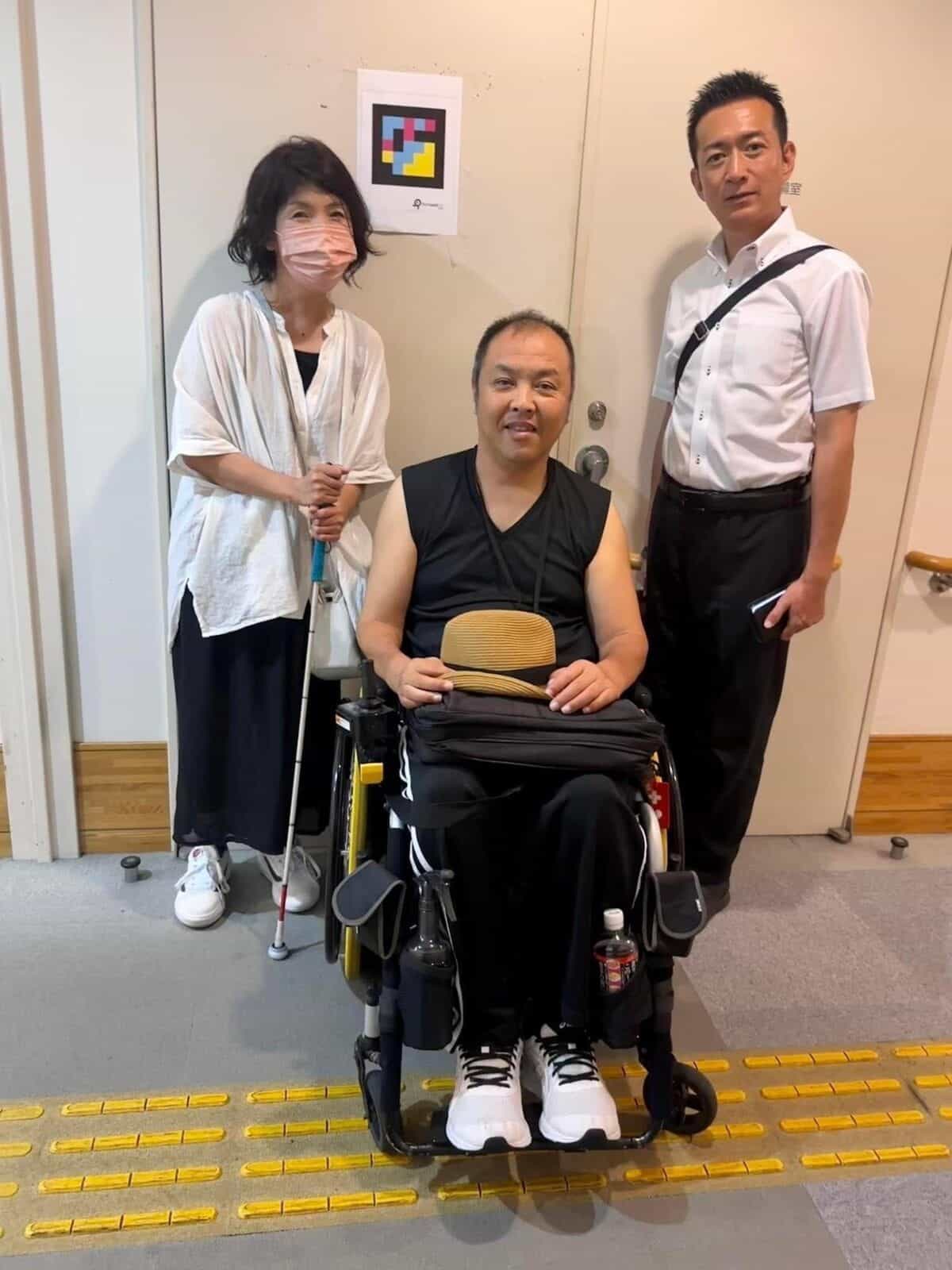 NaviLens 案内板が貼られたアクセシブルトイレのドアの前に立つまゆだまネットフェスタ参加者3名、うち1名は電動車いす