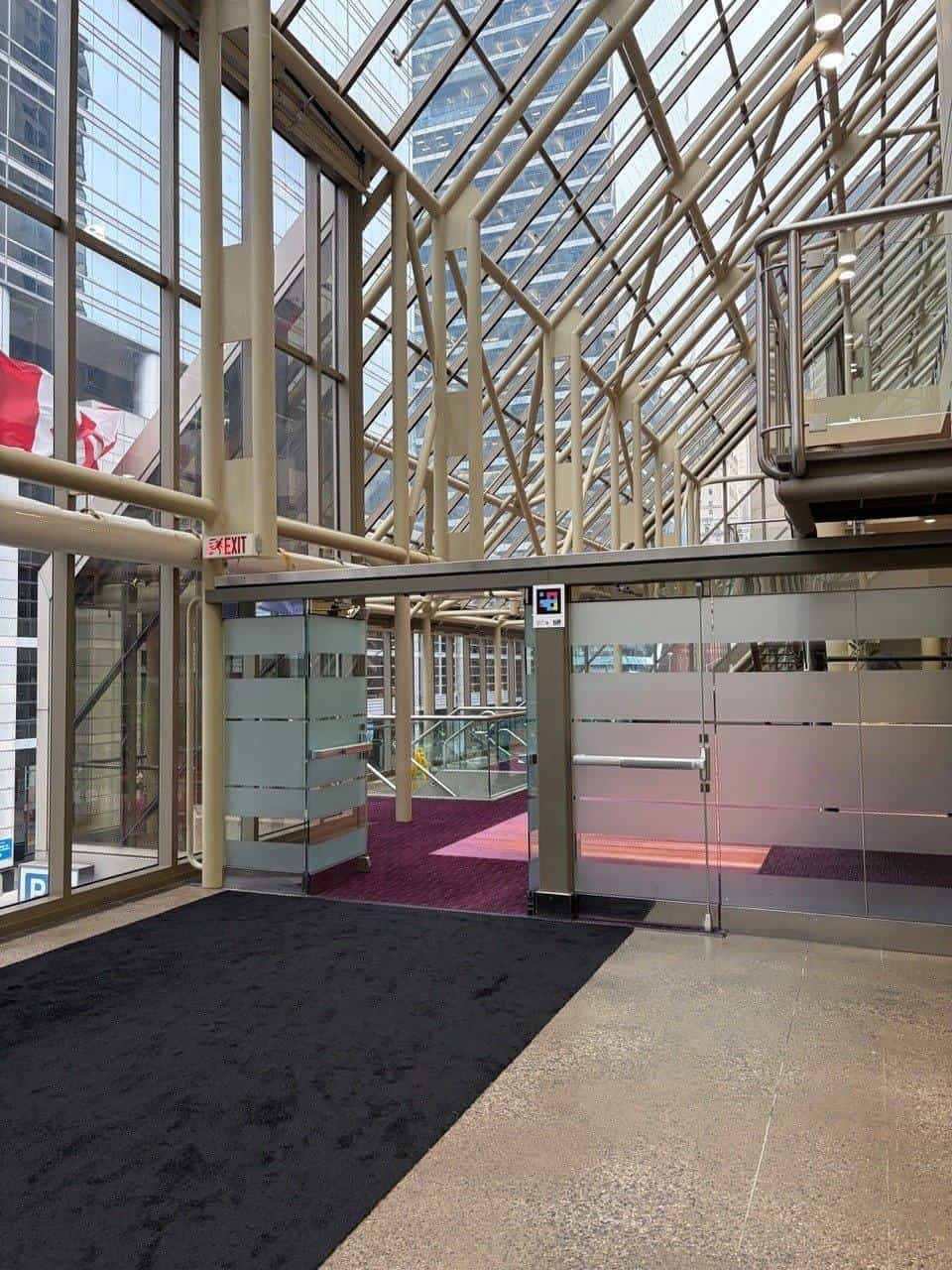 The glassed-in skywalk at the Metro Toronto Convention Centre: pale steel trusses under a glass roof with the Toronto skyline behind. On the central column, next to the glass doors, a NaviLens code with the TIFF logo.