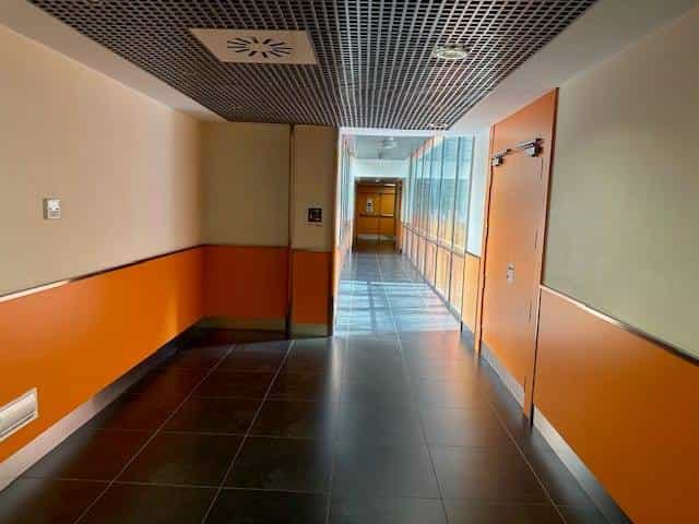 Glazed corridor connecting buildings at CRE ONCE Madrid: orange baseboard on both sides, a continuous window on the right opening onto an interior courtyard and a metal grid ceiling