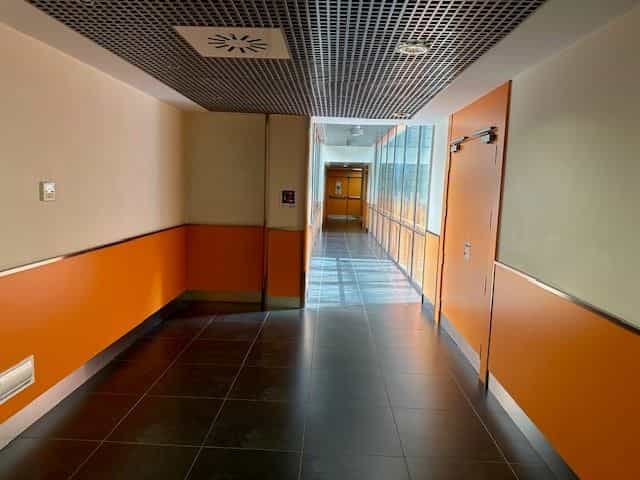 Verglaster Verbindungsgang zwischen Gebäuden im CRE ONCE Madrid: orangefarbener Sockel auf beiden Seiten, durchgehendes Fenster rechts mit Blick auf einen Innenhof und Metallgitterdecke