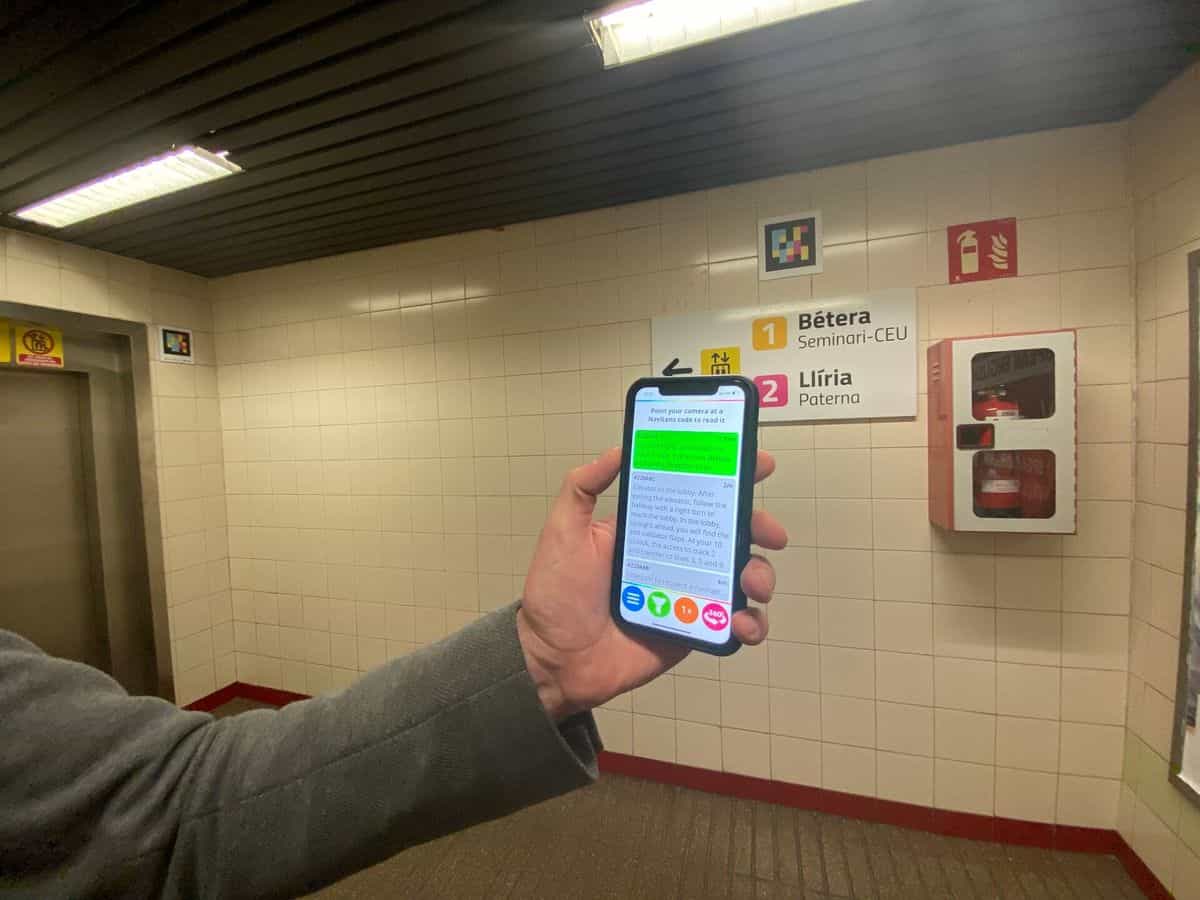 Pasillo blanco con cartel '1 Bétera Seminari-CEU · 2 Llíria Paterna' y, sobre los azulejos, un código NaviLens cuadrado a colores; en primer plano, una mano sostiene un móvil con la app NaviLens en inglés indicando 'On your right, an elevator to track 1, Line 1 direction Bétera and Line 2 direction Llíria' a 1,97 m
