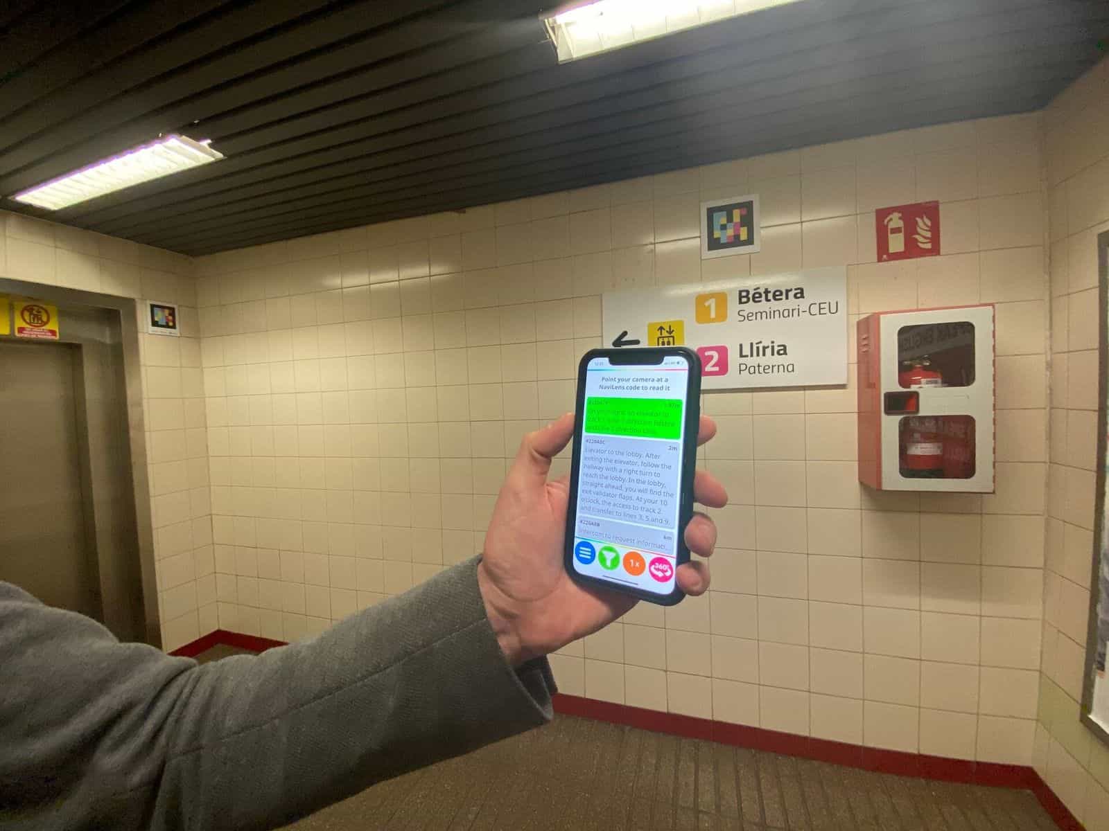 Corridoio bianco con il cartello «1 Bétera Seminari-CEU · 2 Llíria Paterna» e, sulle piastrelle, un codice NaviLens quadrato a colori; in primo piano, una mano tiene un telefono con l’app NaviLens in inglese che indica «On your right, an elevator to track 1, Line 1 direction Bétera and Line 2 direction Llíria» a 1,97 m