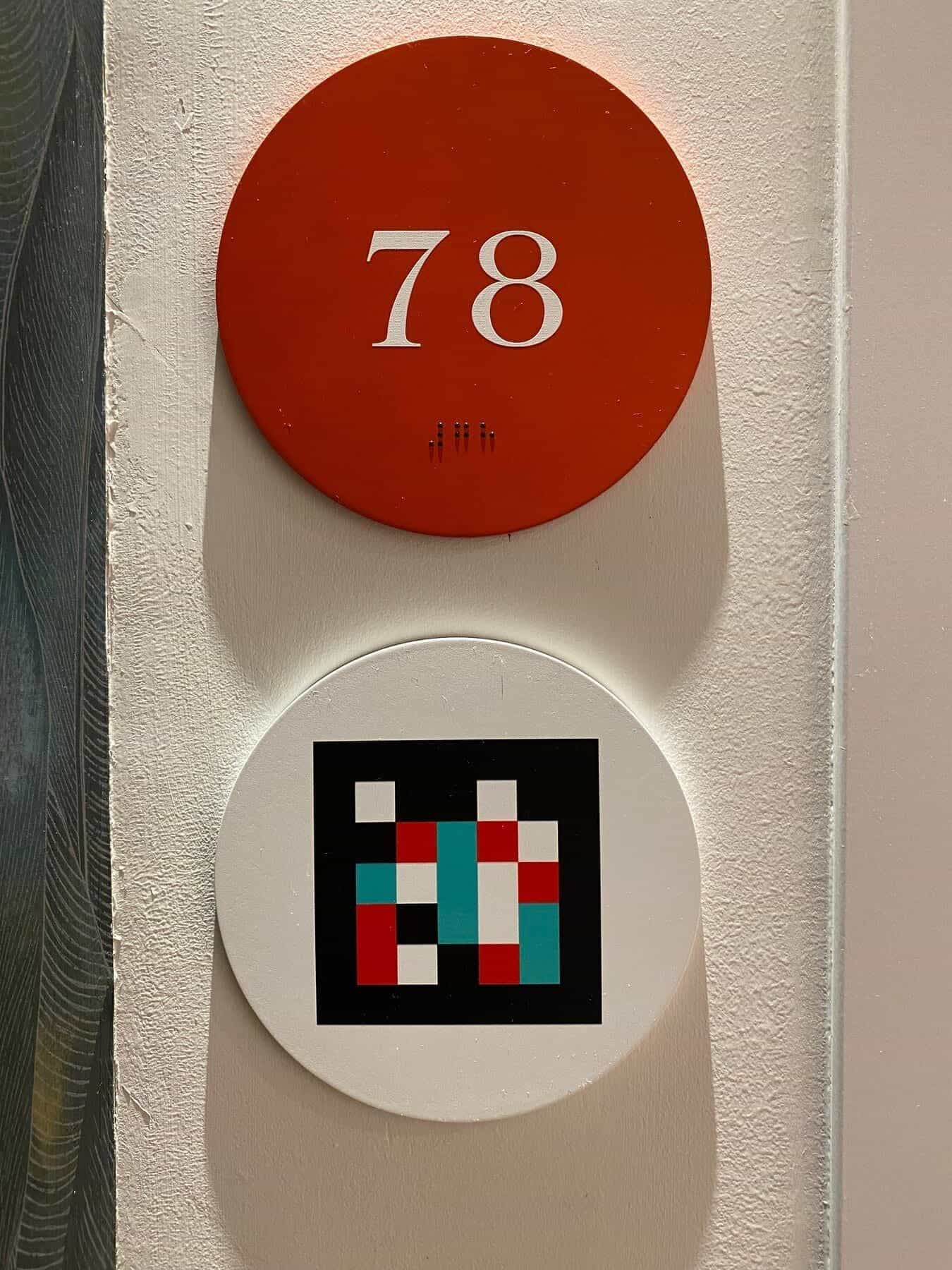 Flur am ibis Styles Sevilla mit orange gepunktetem Teppich: links der Wegweiser mit NaviLens-Code, rechts der Türrahmen von Zimmer 67 mit rotem Disc und NaviLens-Code