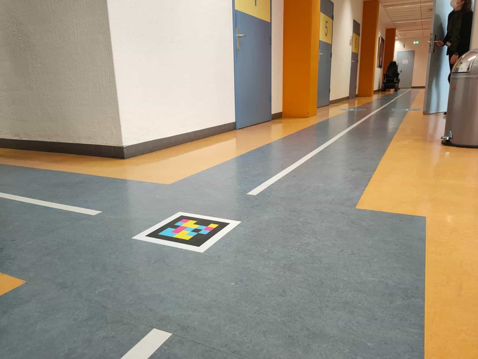 Ambulanzflur des Radboudumc mit grau-blauem Boden und orangen Streifen; gelbe Säulen, blaue Türen mit den Nummern 5 und 6 und im Hintergrund ein Elektrorollstuhl. Im Vordergrund auf dem Boden ein weiß umrahmter NaviLens-Code neben einer Abstandsmarkierung