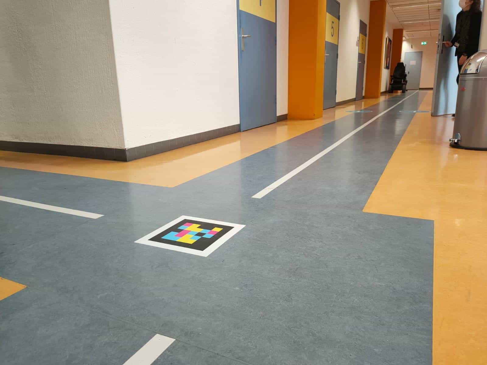 Pasillo de consultas numeradas en el Radboudumc de Nijmegen con códigos NaviLens en el suelo a cada puerta
