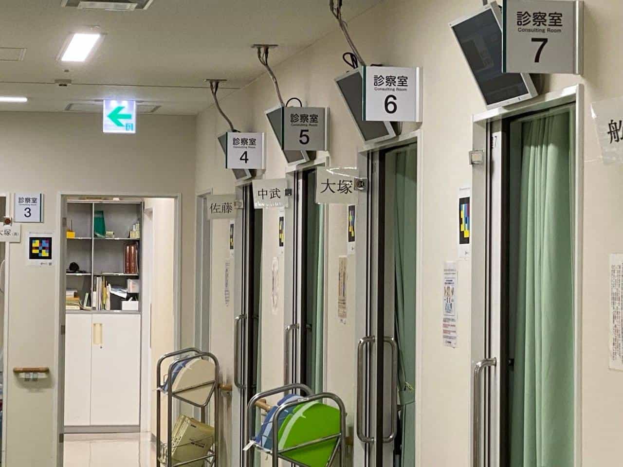 Ophthalmology corridor at Oita University Hospital: sliding doors with green curtains and, above them, the “診察室 / Consulting Room 4, 5, 6, 7” signs in white with a large black number; on each white door frame there is a colour-tile NaviLens code printed on a laminated sheet, and on each door the doctor's name (佐藤・中武・大塚) written in kanji
