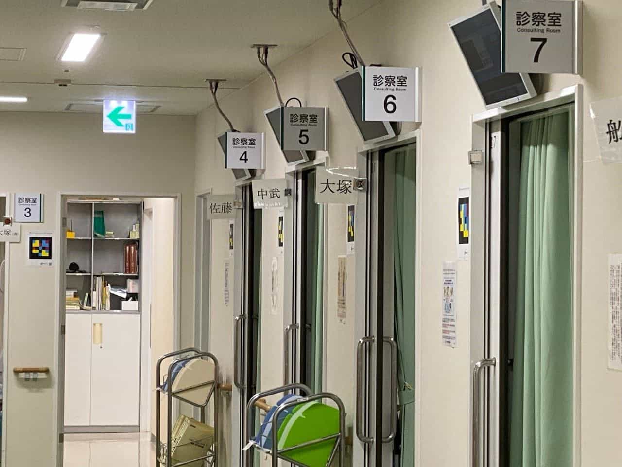 大分大学医学部附属病院 眼科の廊下：緑のカーテンが付いた引き戸の上に「診察室 / Consulting Room 4, 5, 6, 7」と大きな黒字番号の白い表示があり、各扉脇の白い枠にNaviLensのカラータイルコードがラミネートされて貼られ、各扉には佐藤・中武・大塚など医師名が漢字で表示されている