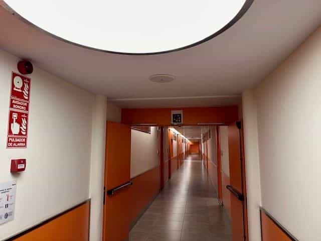 Wohnheimgang des CRE ONCE Madrid in der Perspektive: weiße Wände und orangefarbener Sockel, Zimmertüren in Reihe, ein roter Alarmschalter im Vordergrund und Oberlicht am Ende