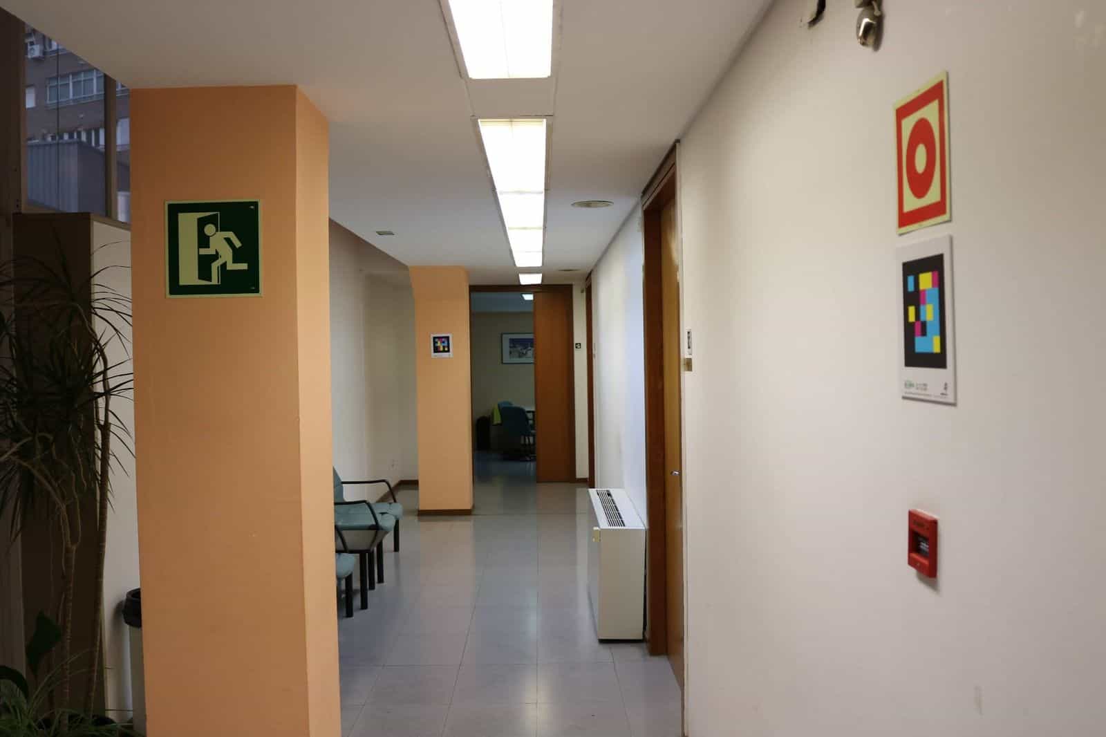 Corridoio interno della sede dell’ONCE Valladolid con pareti color salmone e bianco: a destra, idrante rosso con codice NaviLens quadrato a colori sotto e pulsante di allarme; in fondo, sala d’attesa con poltrone azzurre e un altro codice NaviLens incorniciato sulla parete