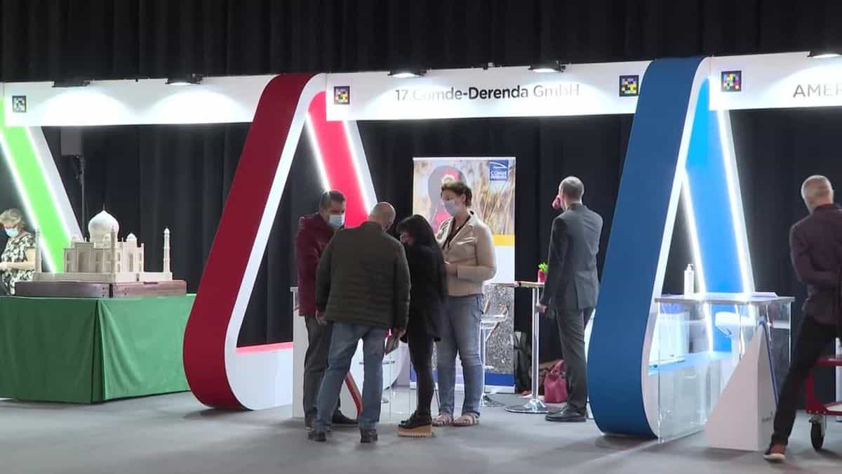 Pasillo central del recinto de Tifloinnova 2022 con grandes arcos en forma de A iluminados con neón verde, rojo y azul; sobre la cabecera blanca de cada stand —Comde-Derenda (17), Ameridian, etc.— aparece el código de colores NaviLens junto al número de stand, permitiendo orientarse de un expositor a otro