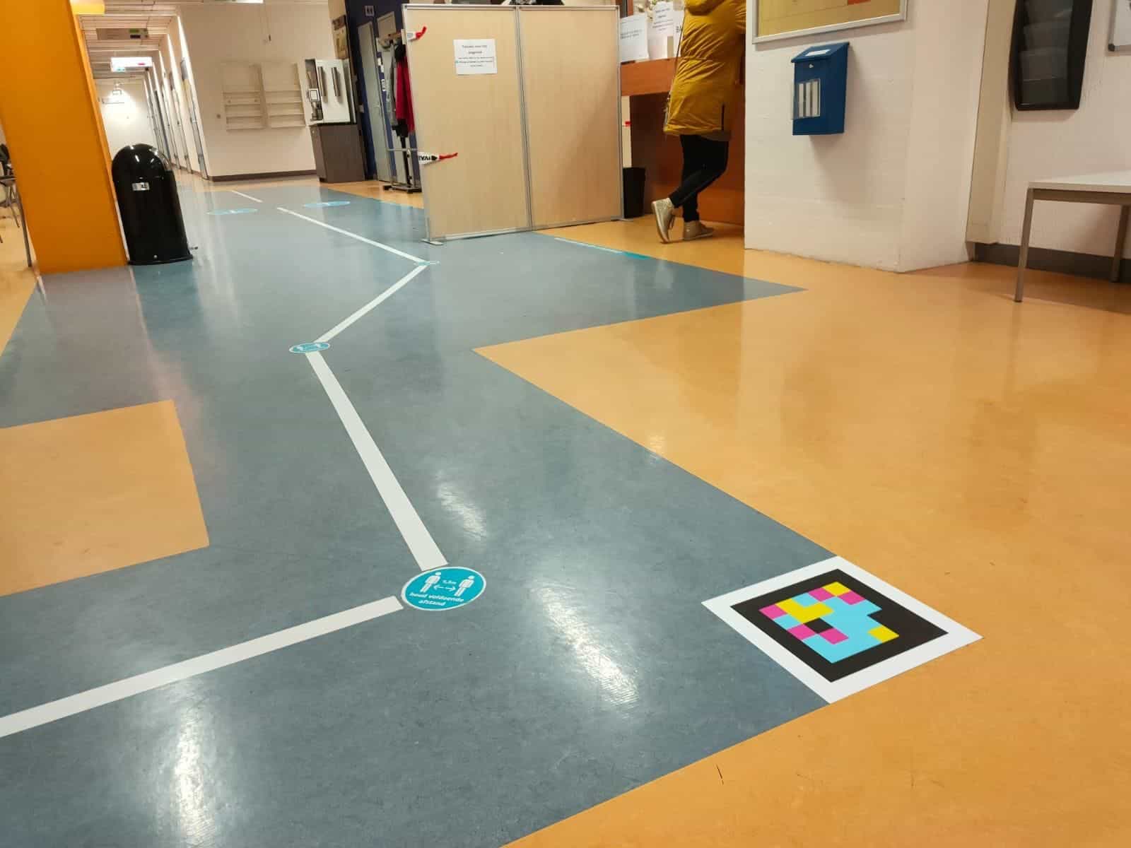 Flur im Radboudumc mit zweifarbigem blau-orangem Boden: eine Zickzack-Route aus weißem Klebeband quert den blauen Streifen, mit runden Aufklebern «Houd voldoende afstand» (Abstand halten). Im Vordergrund auf dem Boden ein weiß umrahmter NaviLens-Code; im Hintergrund betritt eine Person in gelbem Mantel einen Raum