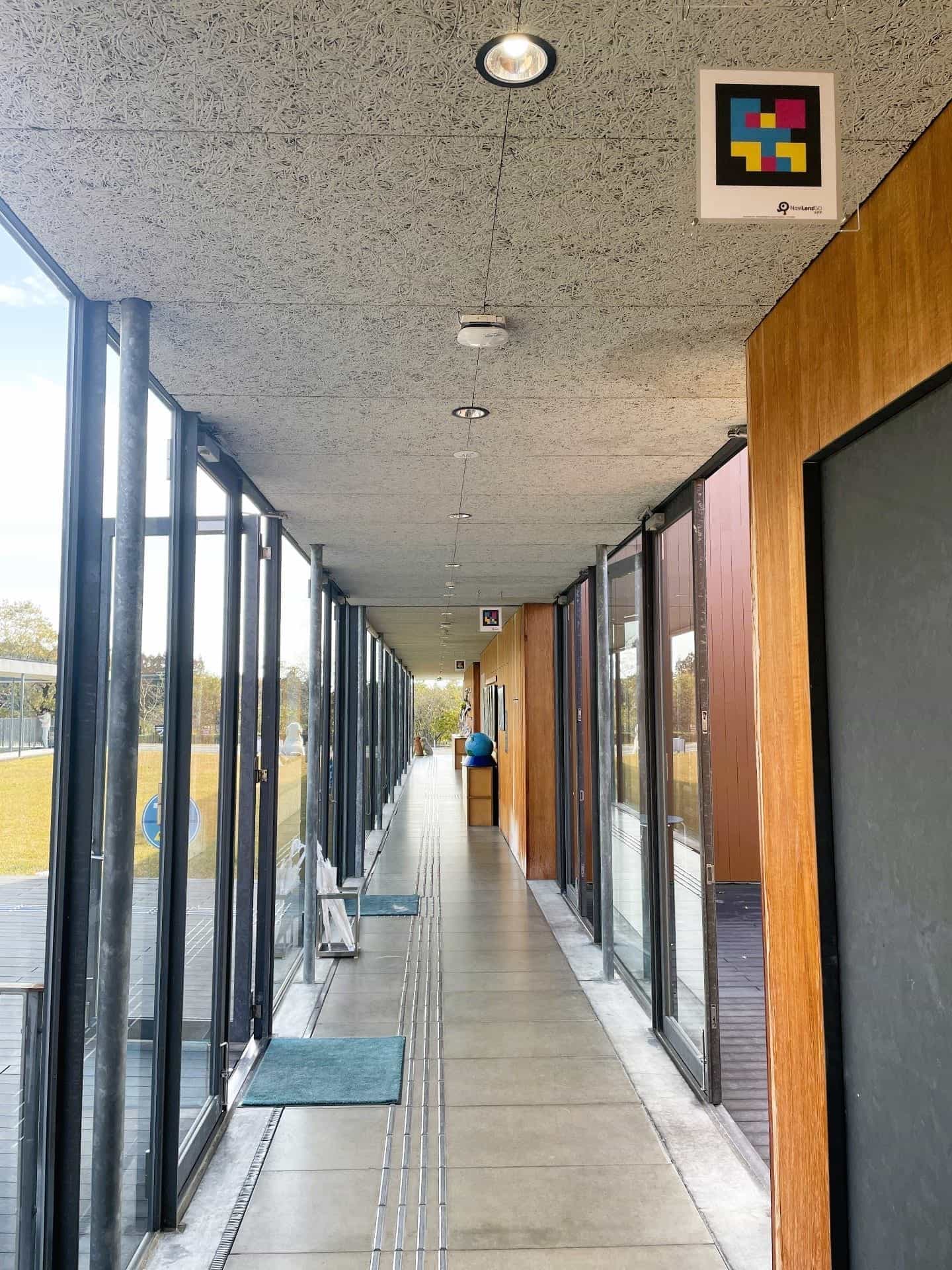 Long glazed corridor at Fuji Harness with continuous tactile paving, wooden walls and a NaviLens GO code on the fiber ceiling at the start of the route