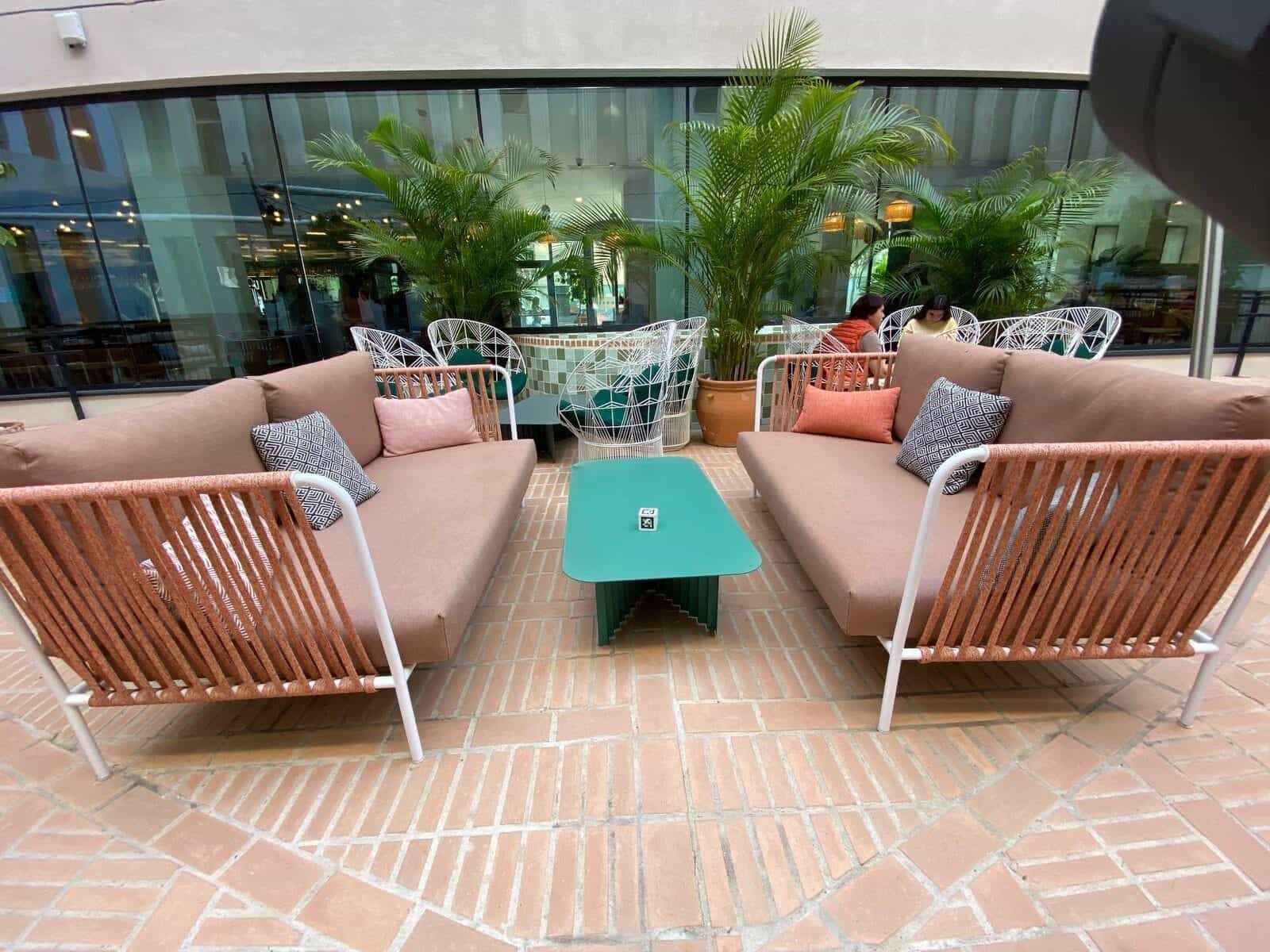 Innenhof am ibis Styles Sevilla mit orangefarbenen Seilsofas, türkisem Tisch mit kleinem NaviLens-Würfel, Palmen und andalusischer Keramik im Hintergrund