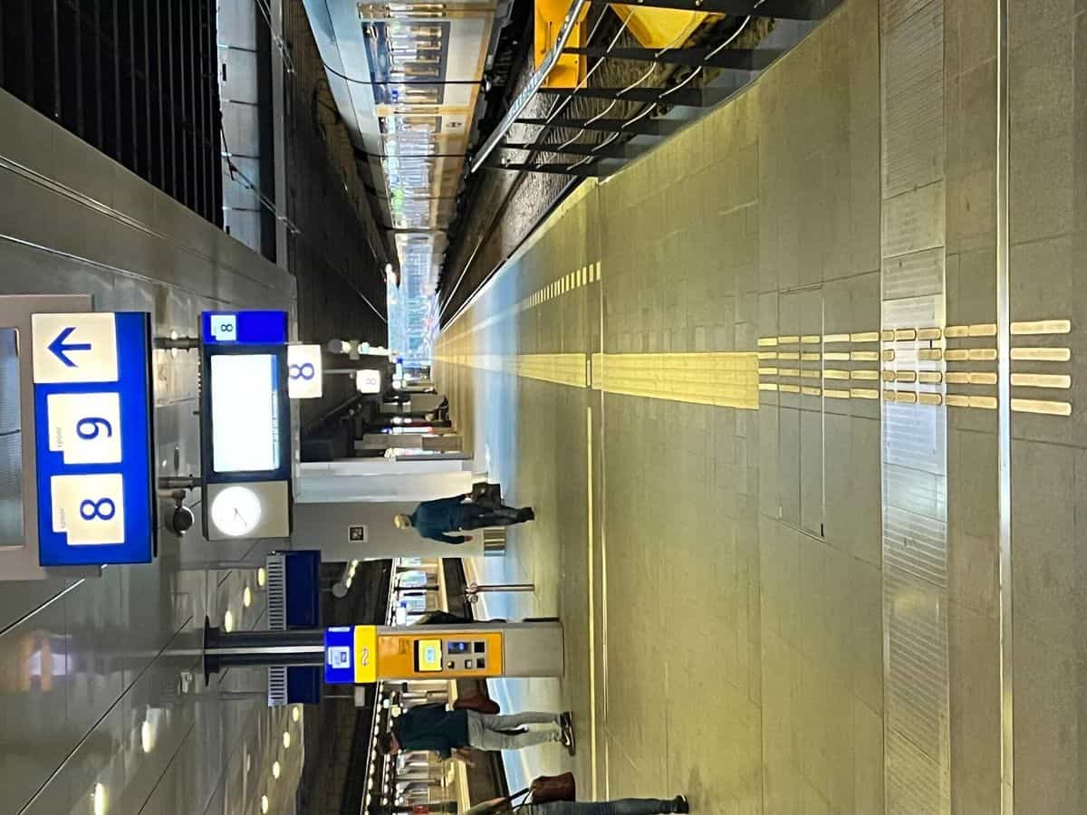 Andén 8/9 de Den Haag Centraal con cartel azul de spoor, máquina amarilla NS y línea-guía táctil de doble franja recorriendo la longitud del perron