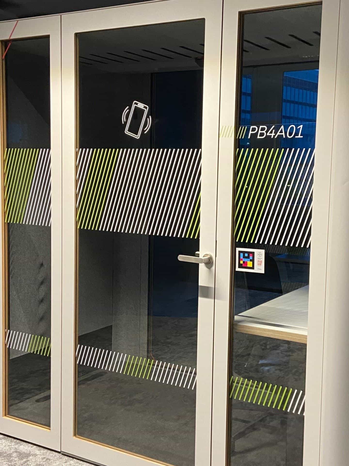 Phone booth PB4A01 della sede Bayer di Leverkusen — cartello NaviLens con croce Bayer sulla porta a vetri, accanto al pittogramma del telefono e all’insegnatura obliqua del booth