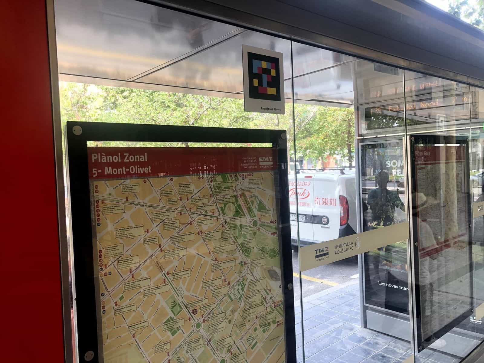 Interno della pensilina con il «Plànol Zonal 5 – Mont-Olivet» a sinistra e, sopra, un codice NaviLens quadrato a colori con il testo «Escaneja amb NaviLens GO»; in fondo, il pannello AVM mostra «Est. del Nord 2 m.»