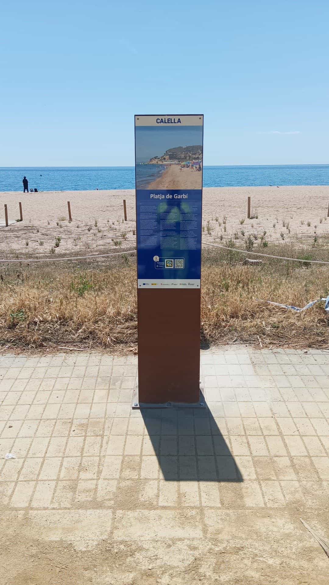 Tótem azul CALELLA con foto de la Platja de Garbí y código NaviLens instalado en la arena junto al paseo