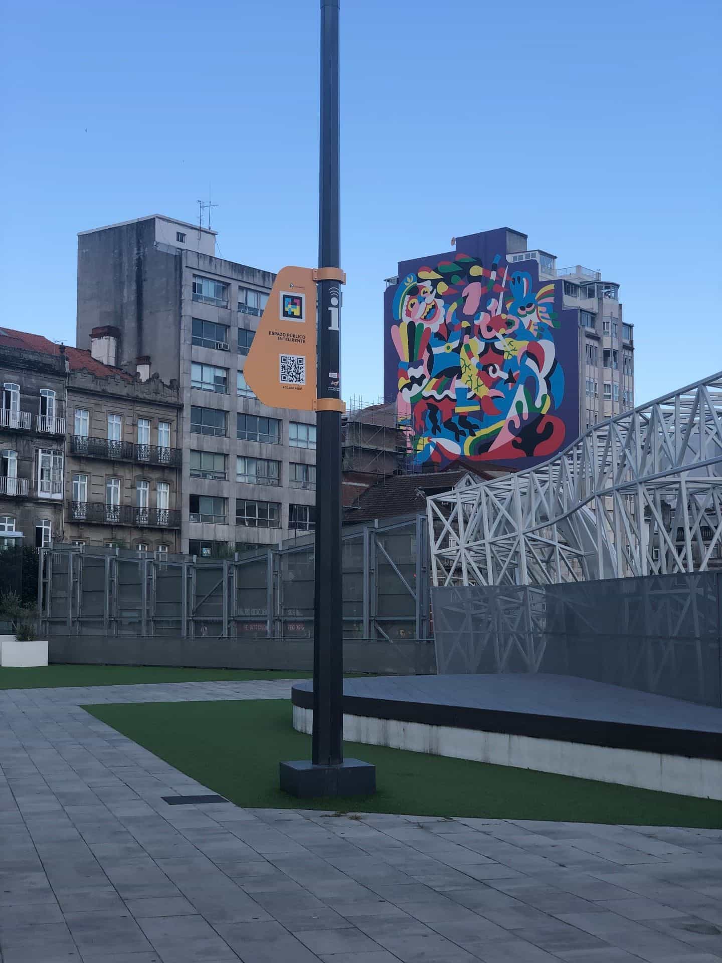 Piazza umanizzata in quota con pedana di legno, erba sintetica e pavimentazione grigia, attraversata da un palo di smart city nero con una targa arancione dell’Espazo Público Intelixente che mostra il codice NaviLens GO e il QR «Accede aquí»; in fondo, l’insieme delle coperture metalliche bianche della stazione di Vialia e un grande murale multicolore su una facciata urbana