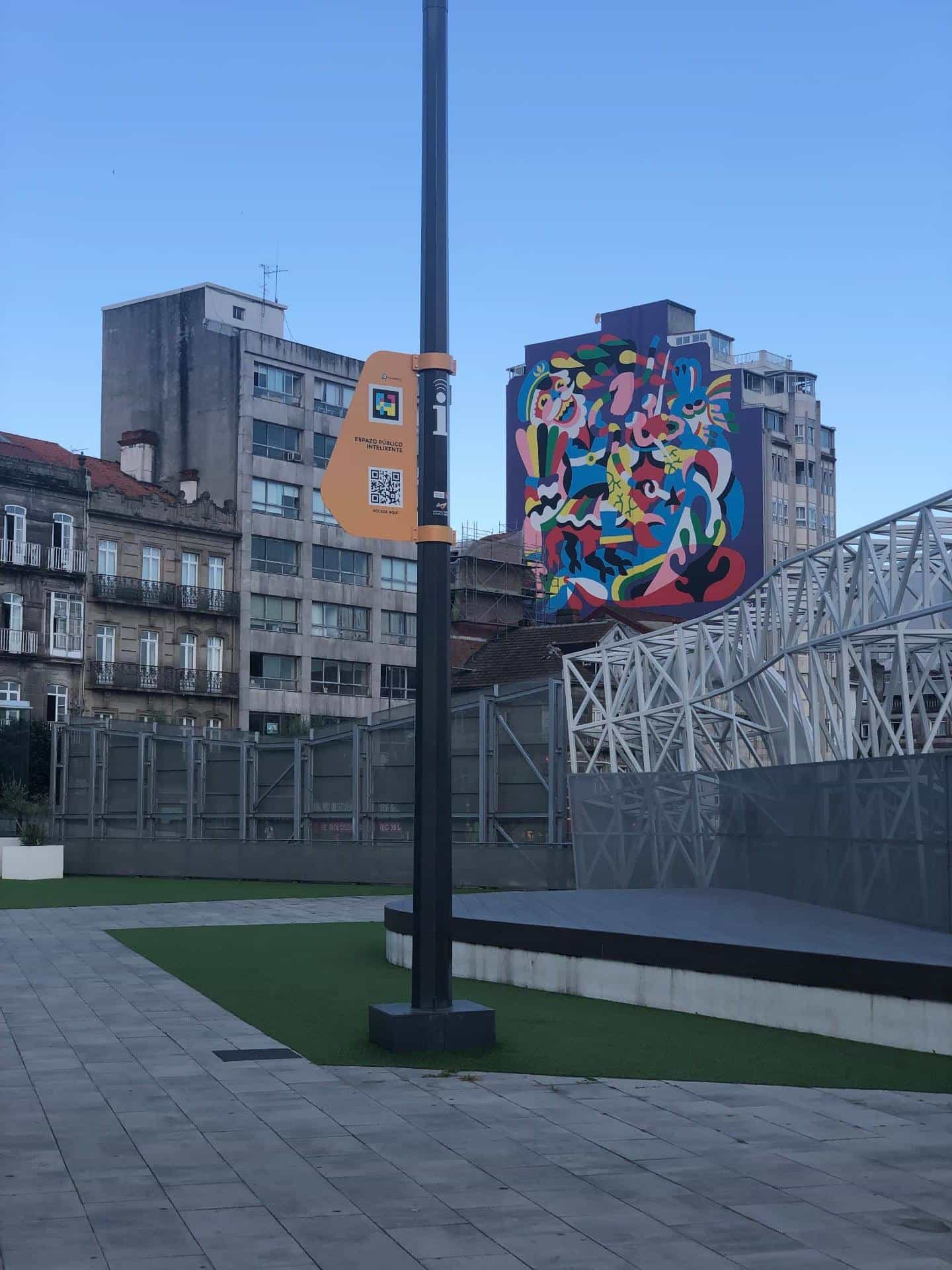 Elevated humanised plaza with wooden decking, artificial turf and grey paving, crossed by a black smart-city post with an orange Smart Public Space plate showing the NaviLens GO code and 'Access here' QR; in the background, the white metal canopies of Vialia station and a large multicoloured mural on an urban facade