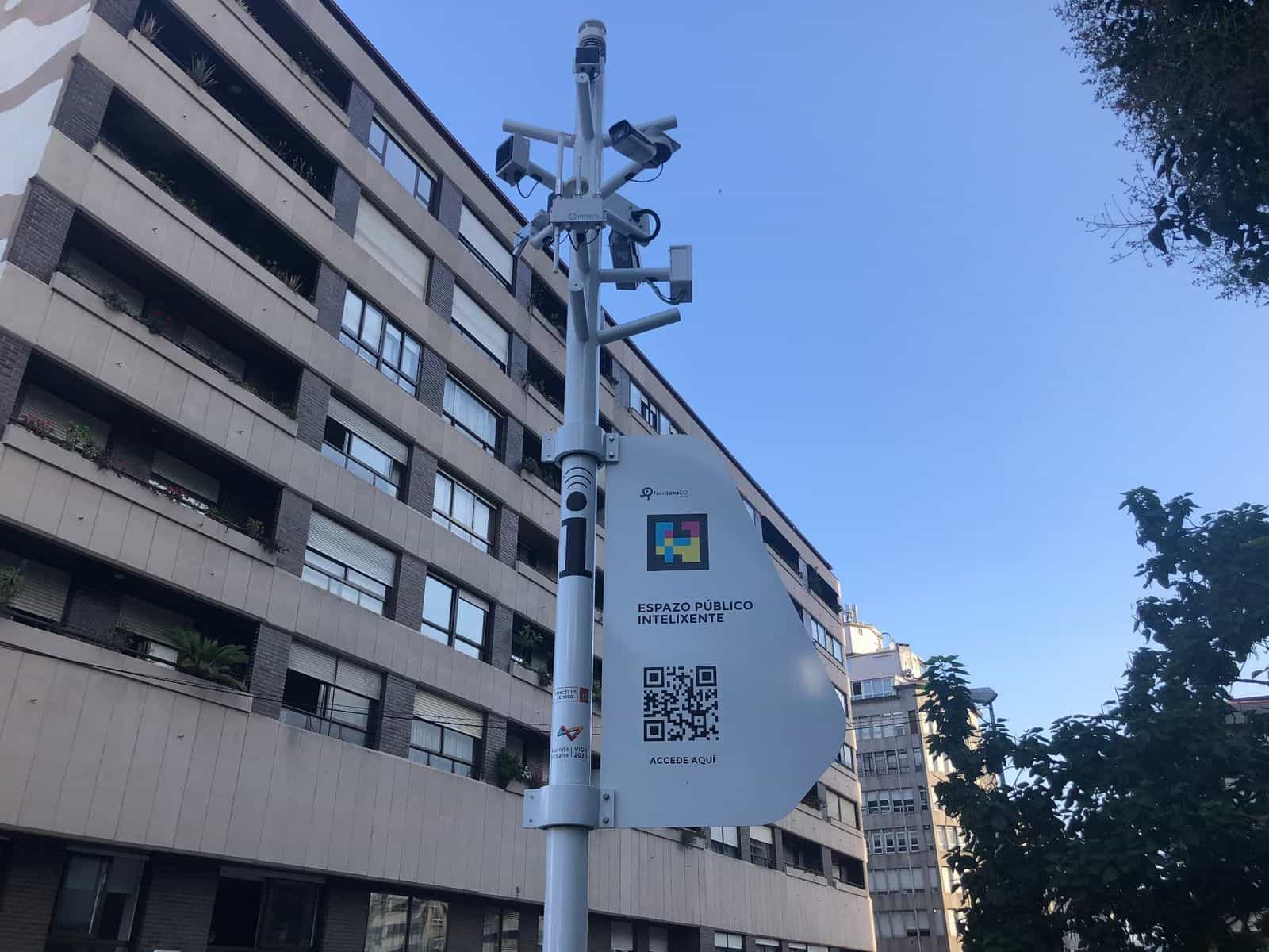 Veduta dal basso di un palo bianco di smart city con diverse telecamere e sensori IOTSENS su fondo di cielo azzurro ed edificio residenziale; sotto le telecamere, una targa bianca curva con il logo NaviLens GO, un codice NaviLens quadrato a colori e un QR «Espazo Público Intelixente · Accede aquí» con i loghi del Comune di Vigo e dell’Axenda Urbana Vigo 2030