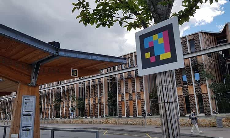 Bus shelter pole with a large multicolour NaviLens code, wooden façade in background.
