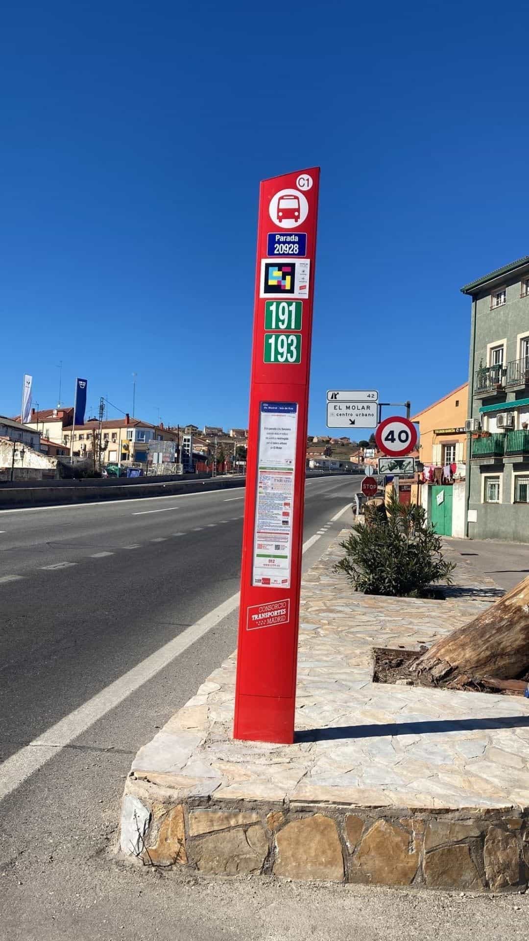 Cartel «Escanea con / Scan with» con un código NaviLens cuadrado a colores, iconos de las apps NaviLens y NaviLens GO, logo del CRTM y logo de Clear Channel pegado sobre el cristal de una marquesina