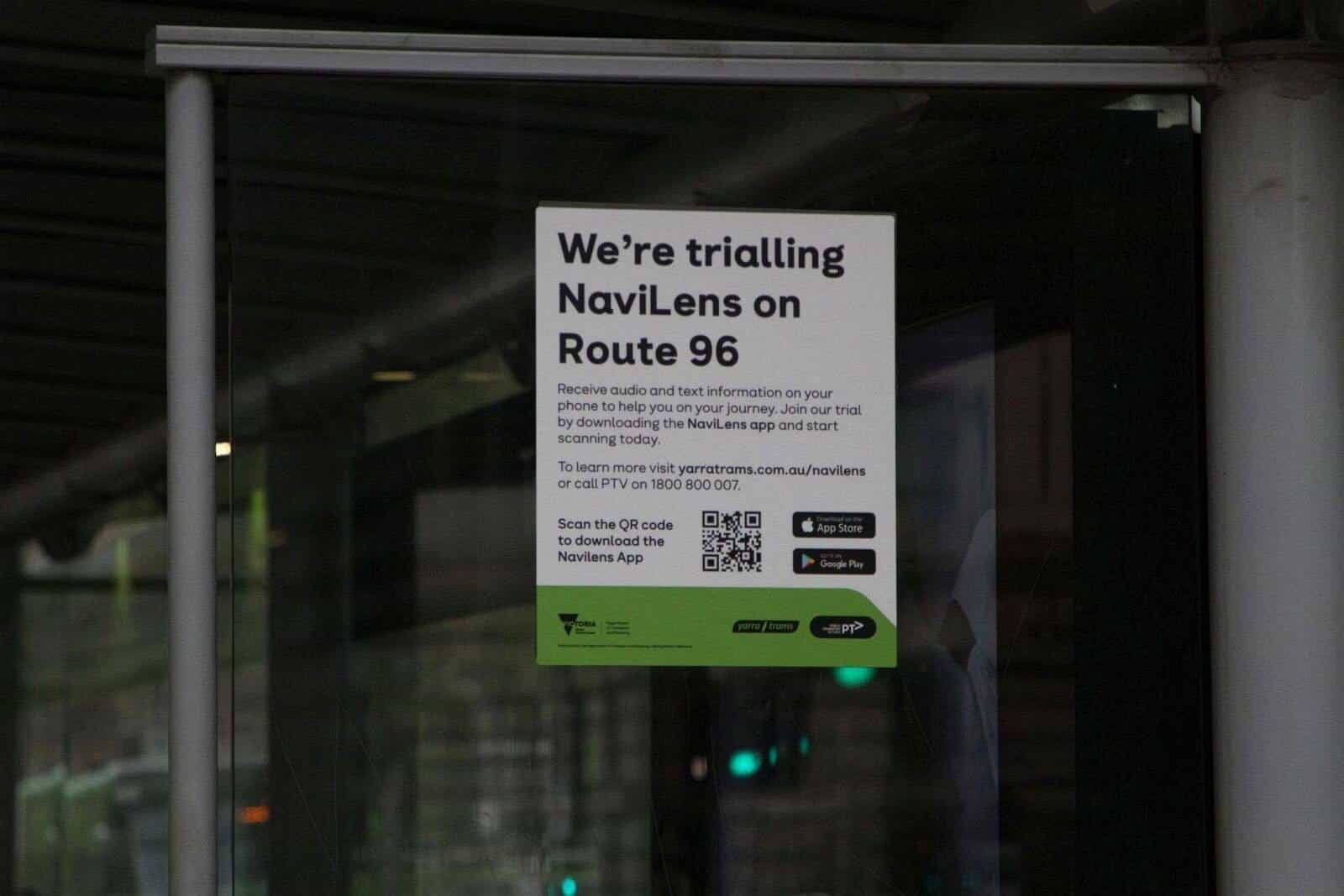 Official poster taped to the inside glass of a Yarra Trams shelter: “We’re trialling NaviLens on Route 96 — Receive audio and text information on your phone…”, with a QR code to download the app, App Store and Google Play icons and a green band with the Victoria State Government, Yarra Trams and PTV logos