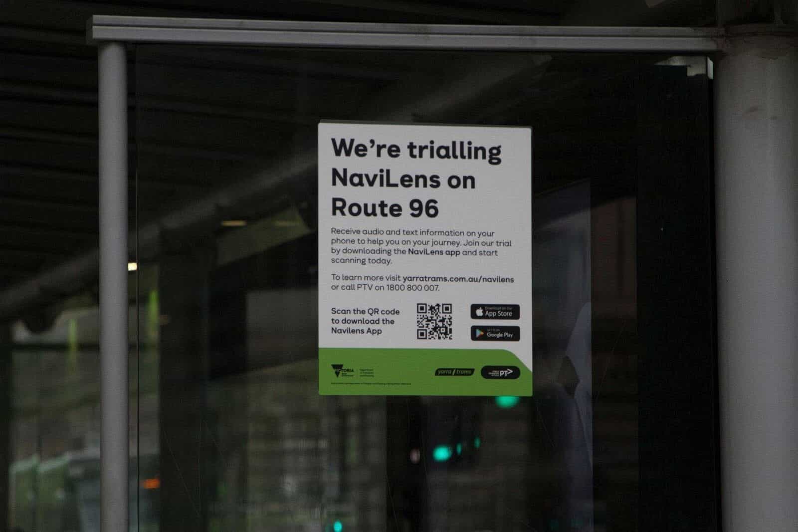 ヤラ・トラムの停留所ガラスの内側に貼られた公式ポスター：「We’re trialling NaviLens on Route 96 — Receive audio and text information on your phone…」、アプリ DL の QR、App Store・Google Play アイコン、緑帯に Victoria State Government・ヤラ・トラム・PTV の各ロゴ