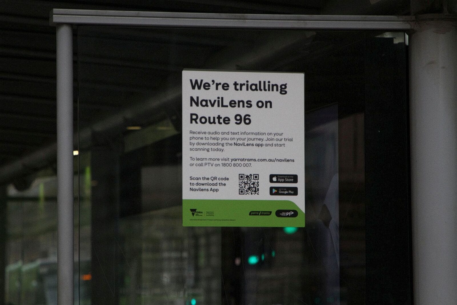 Official poster taped to the inside glass of a Yarra Trams shelter: «We’re trialling NaviLens on Route 96 — Receive audio and text information on your phone…», with a QR code to download the app, App Store and Google Play icons and a green band with the Victoria State Government, Yarra Trams and PTV logos