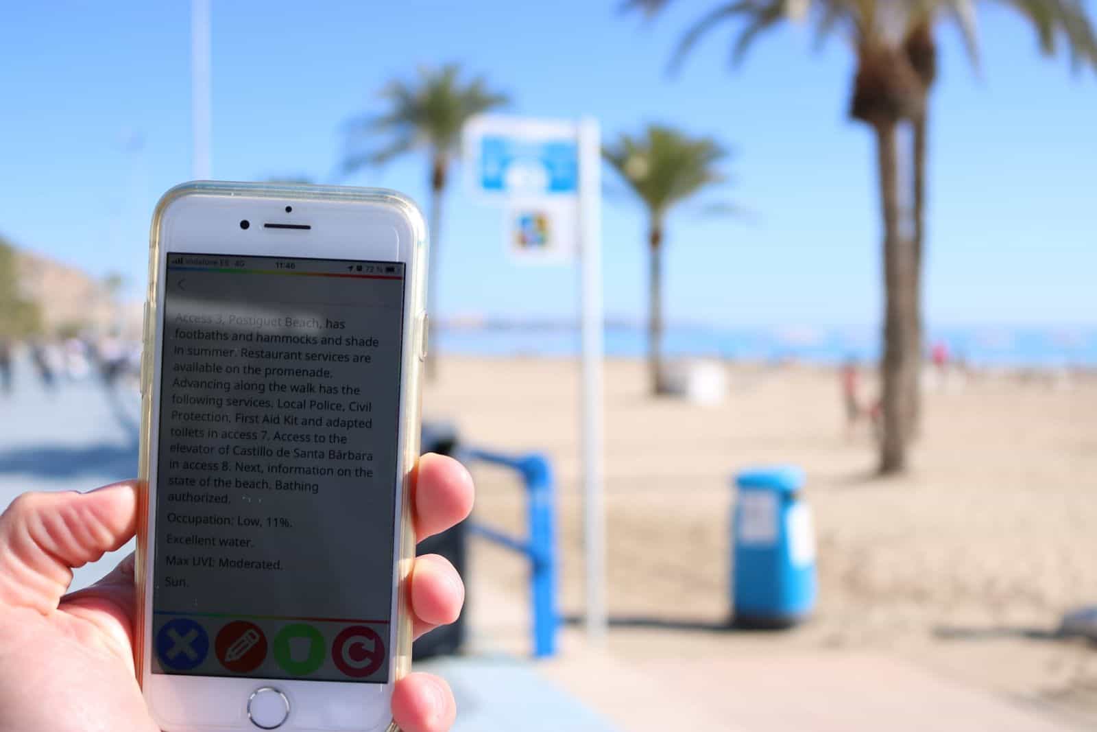 Handy liest einen NaviLens-Code am Zugang 3 des Strandes Postiguet mit englischer Information zu Service, Auslastung, Wasser und UV-Index