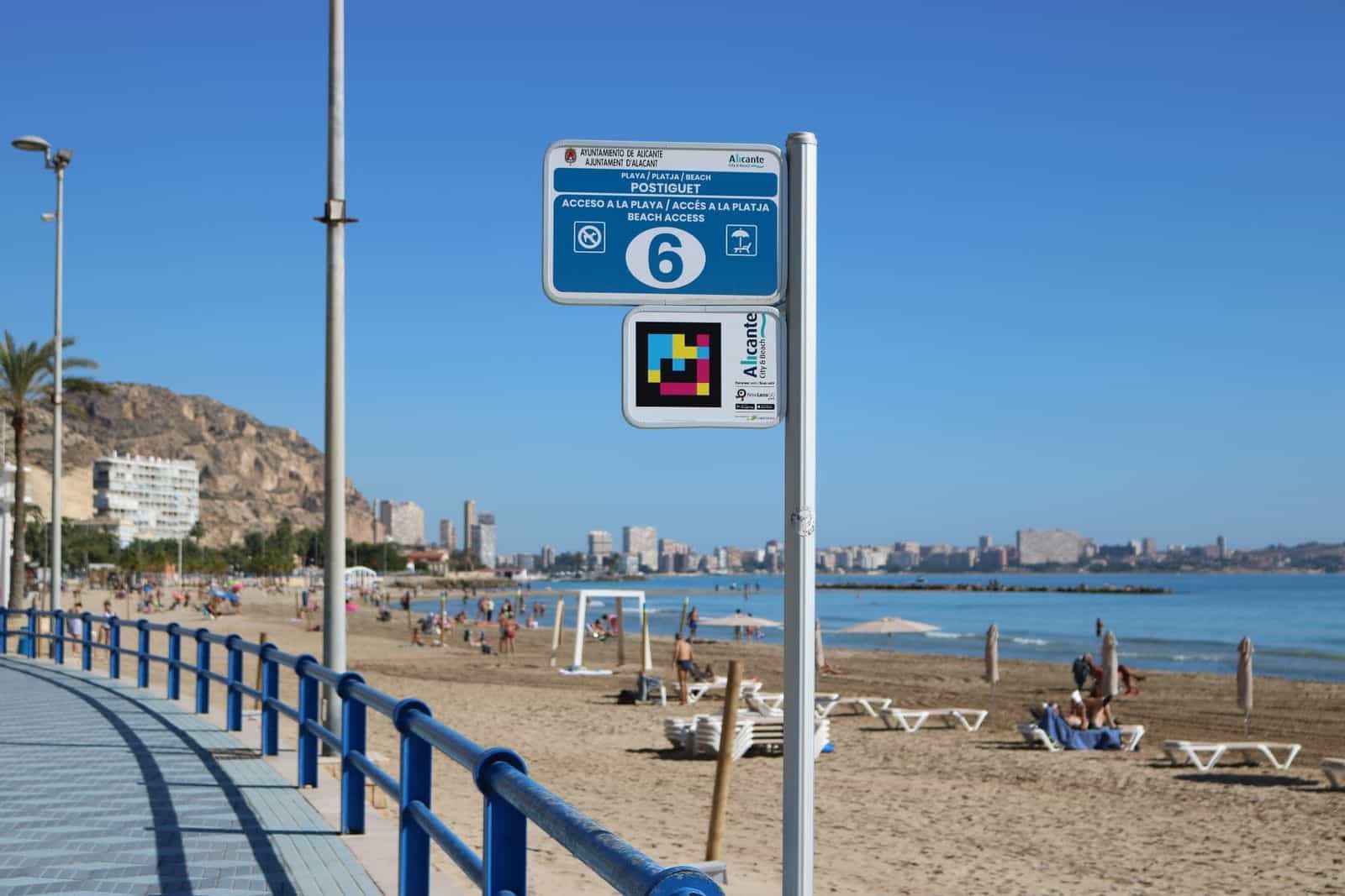 Informationspfosten am Zugang 6 zum Strand Postiguet (Alicante) mit dem NaviLens-Code der Stadt Alicante und der Stadt im Hintergrund