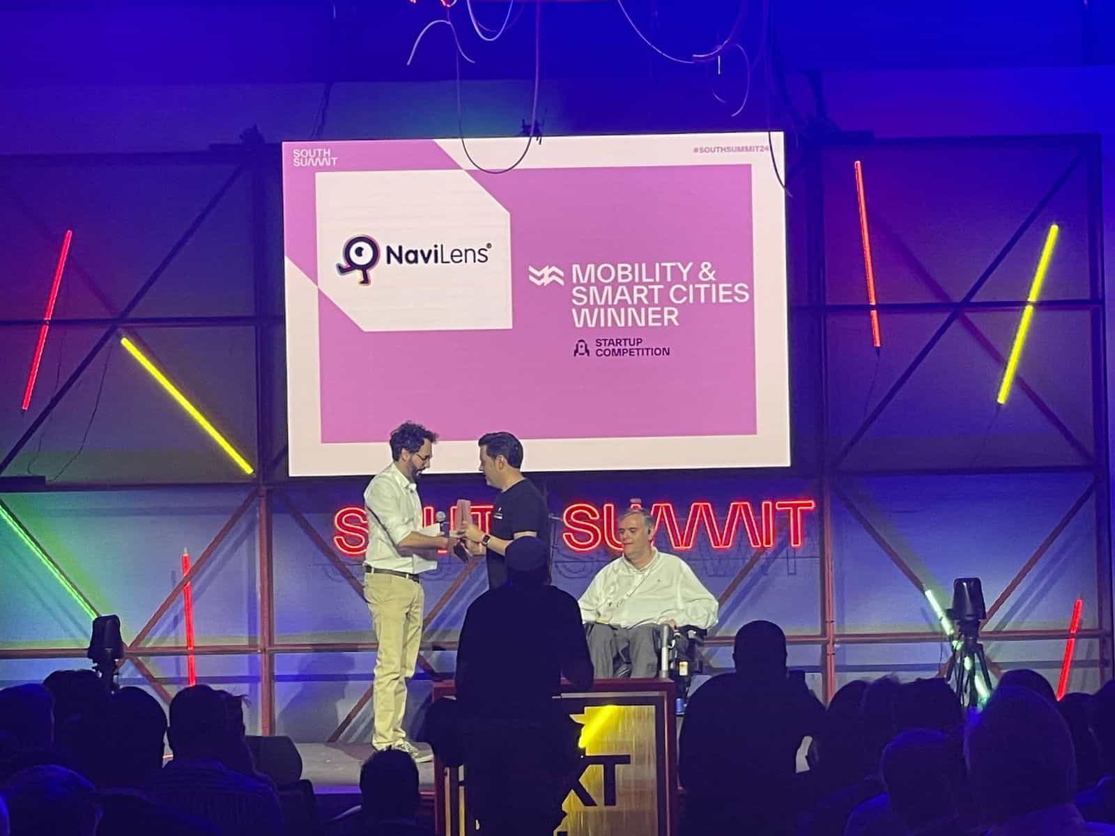 Palco del South Summit Madrid 2024: grande schermo con la slide «NaviLens · Mobility & Smart Cities Winner — Startup Competition #SOUTHSUMMIT24» su fondo rosa; sotto, sul palco illuminato di azzurro e rosso, due rappresentanti si scambiano il trofeo del premio davanti al pubblico e all’insegna al neon rossa «South Summit»