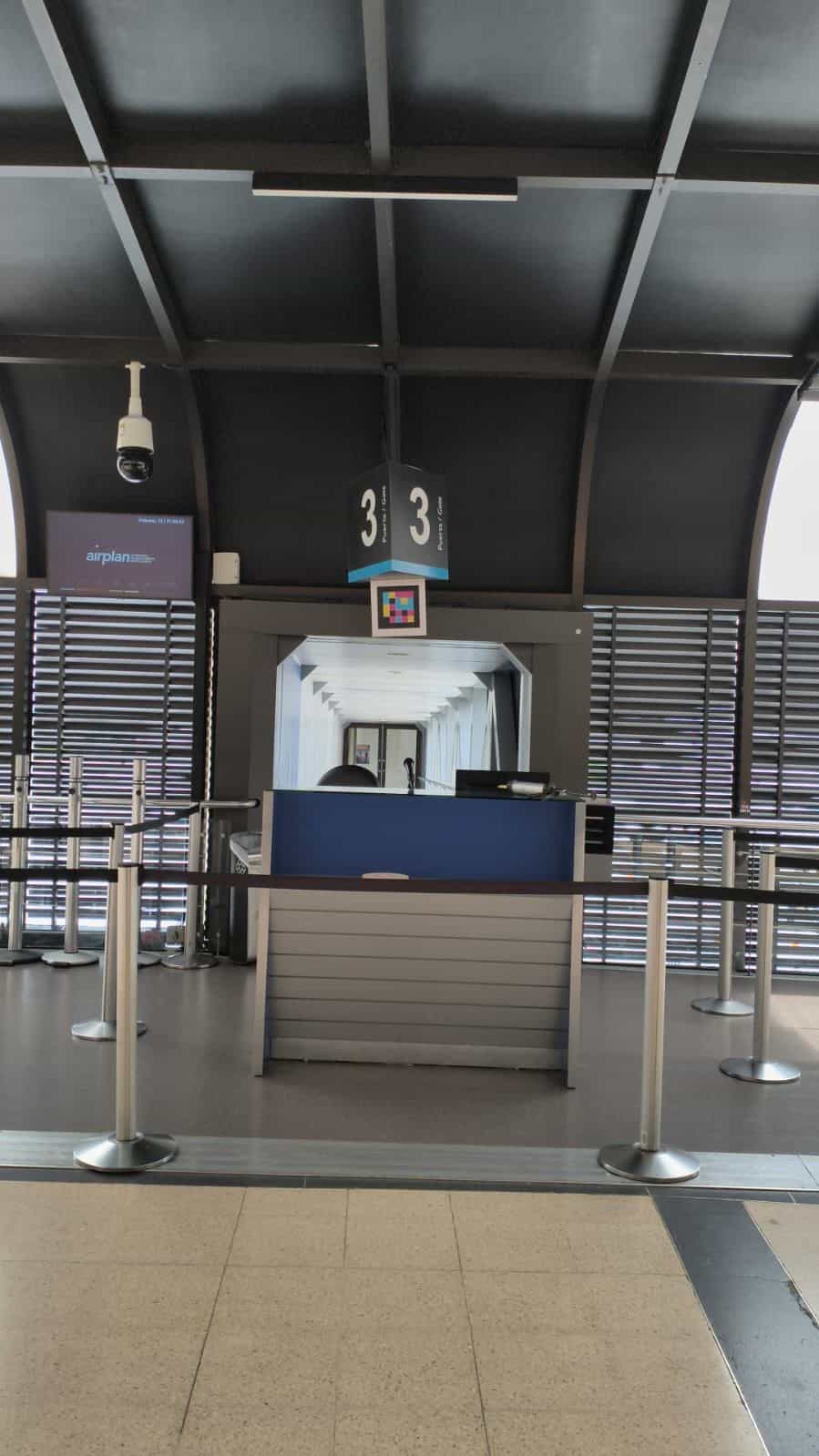 Gate 3 am Flughafen José María Córdova: schwarzes Hängeschild mit weißem Würfel «3 Puerta · Gate» und direkt darunter quadratischer NaviLens-Code in Cyan, Magenta, Gelb und Rot, weiß gerahmt; leerer marineblauer Tresen mit verchromten Pfosten, seitlicher Bildschirm mit Airplan-Fahrplan und im Hintergrund der verglaste Tunnel der Fluggastbrücke