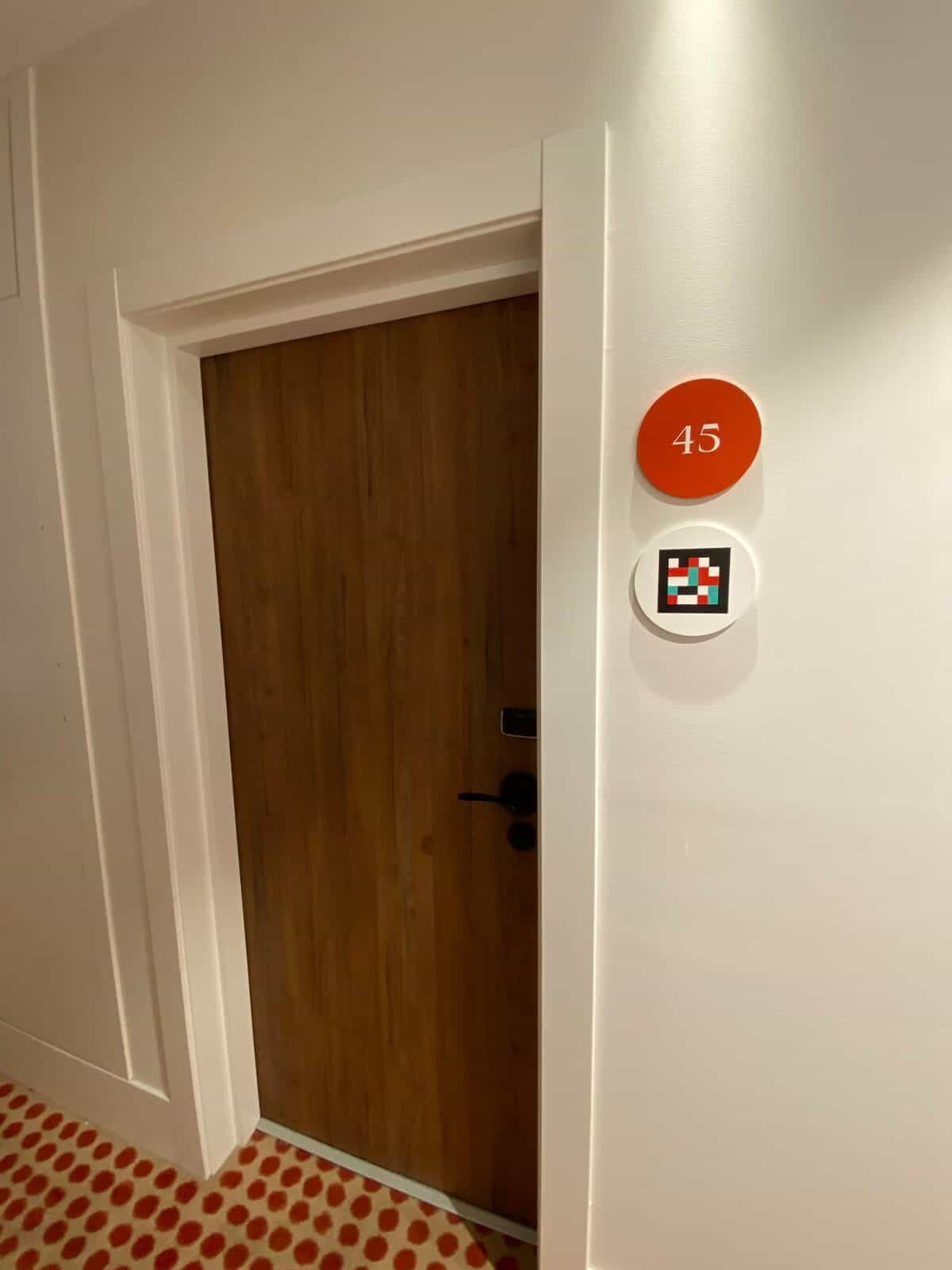 Zimmer 45 am ibis Styles Sevilla: Holztür mit schwarzer Klinke und am weißen Türrahmen ein oranger Disc mit „45“ und darunter ein NaviLens-Code